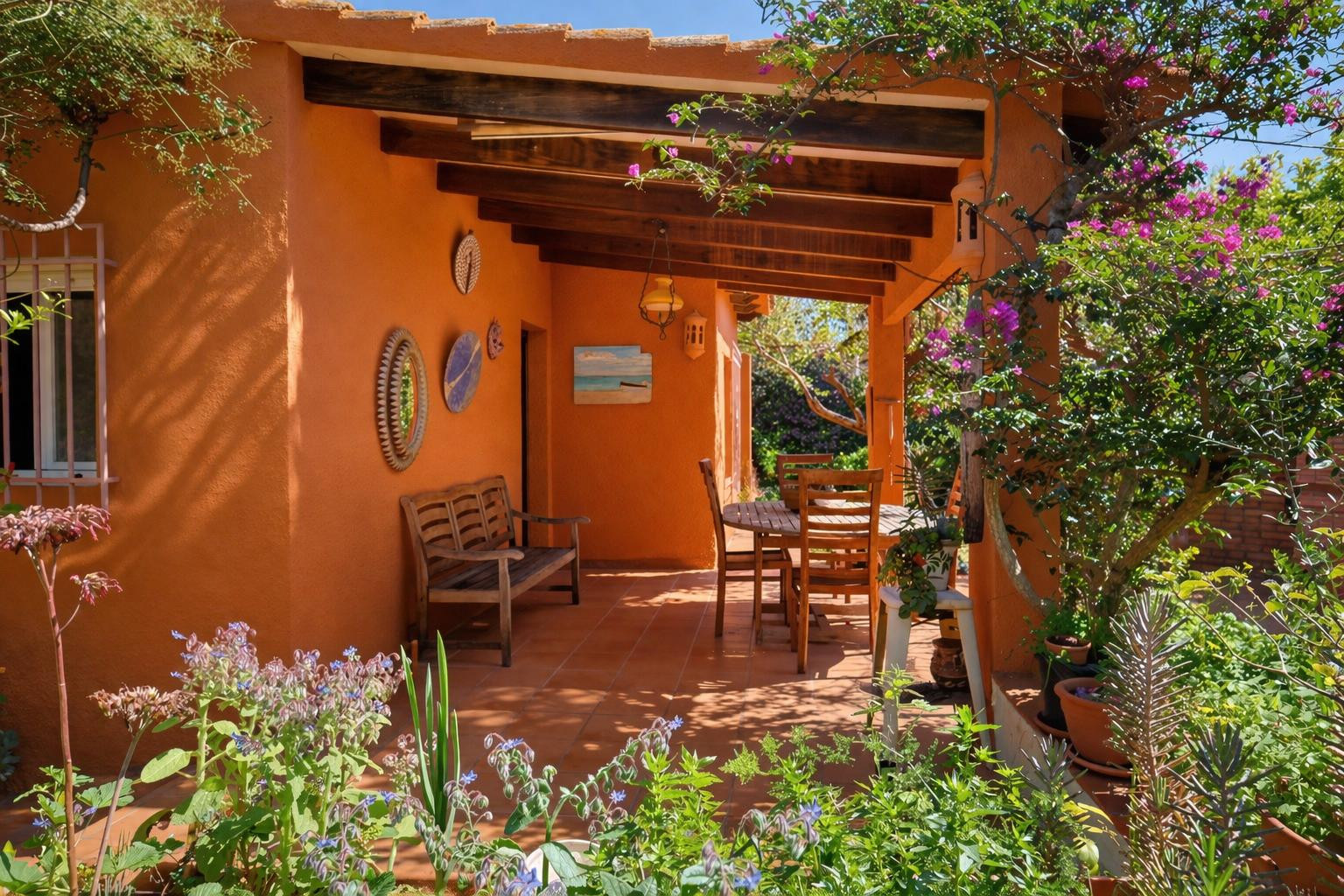 Terrasse couverte avec poutres en bois apparentes, sol en terre cuite et mobilier de jardin, entourée d'une végétation méditerranéenne luxuriante.