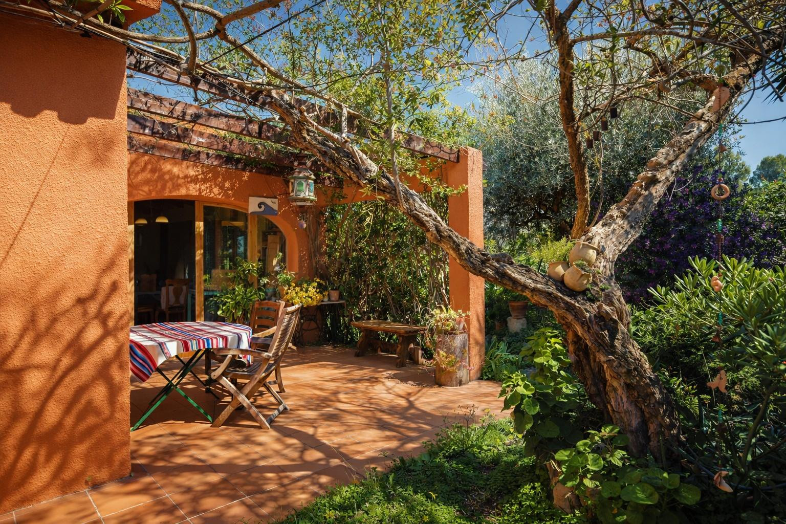 Terrasse extérieure avec sol en terre cuite, pergola en bois et végétation dense offrant un ombrage naturel sur l'espace repas.