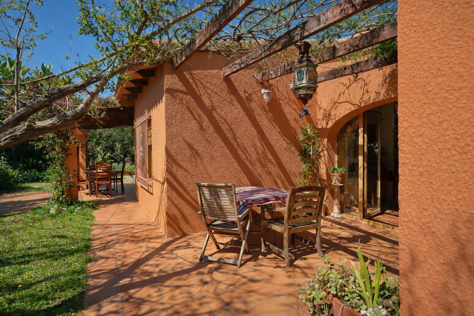 Terrasse extérieure avec sol en terre cuite, pergola en bois végétalisée et mobilier de jardin rustique devant une façade aux tons ocre.