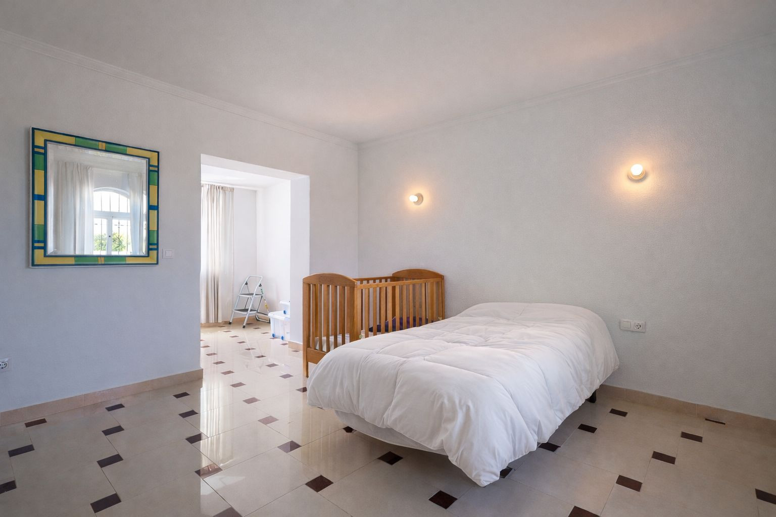 Chambre lumineuse avec sol en grès décoré, lit simple et berceau en bois. Comprend des appliques murales et un accès à une pièce adjacente baignée de lumière.