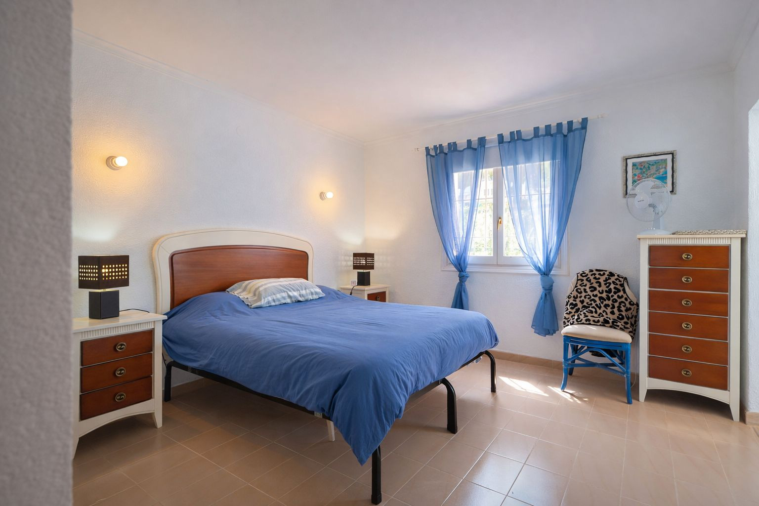 Chambre lumineuse avec sol en grès, lit double avec tête de lit en bois et mobilier assorti. Comprend une commode haute et une fenêtre avec des rideaux bleus.