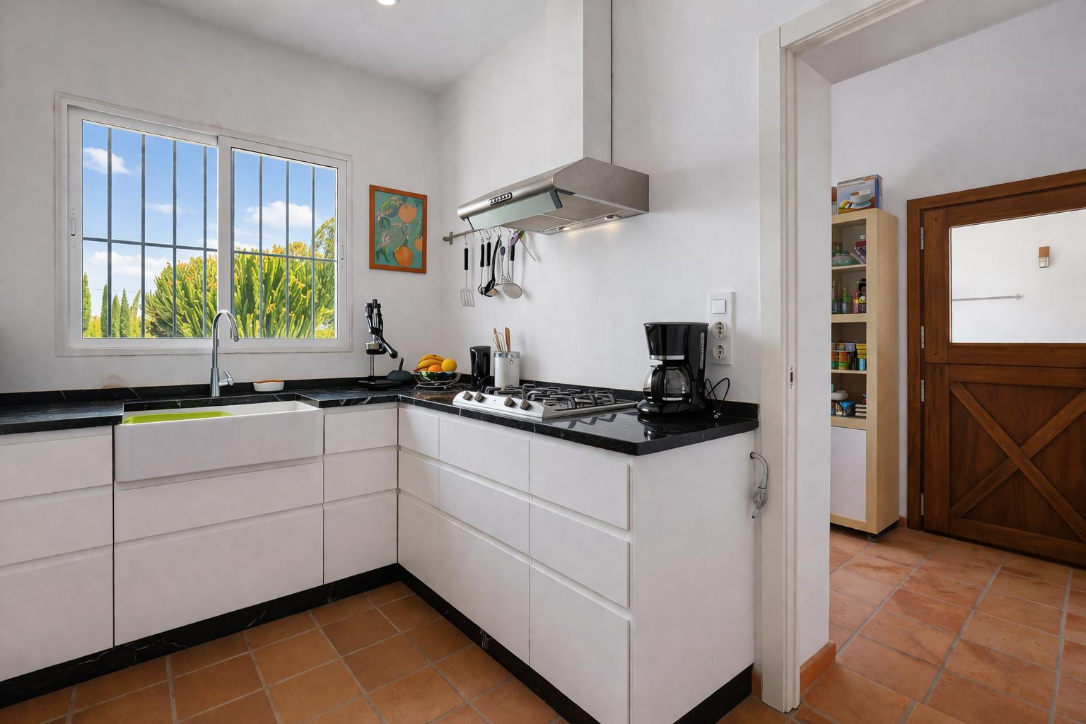Cuisine fonctionnelle avec plan de travail en pierre noire, meubles blancs et évier timbre d'office. Le sol en terre cuite et la lumière naturelle créent un espace clair.