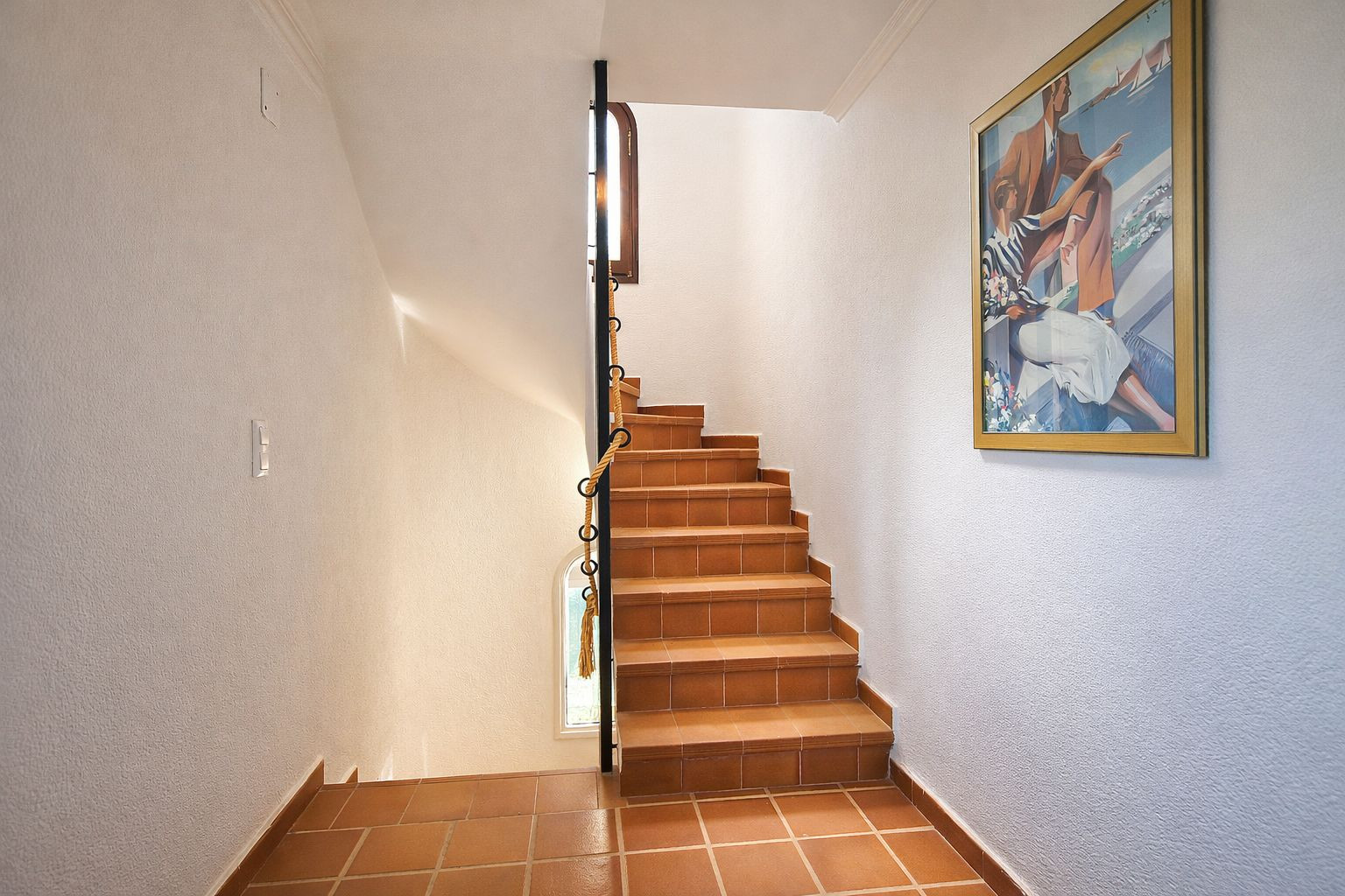 Escalier en terre cuite avec rampe en fer forgé et détail de corde, murs blancs texturés et éclairage naturel provenant d'une petite fenêtre.