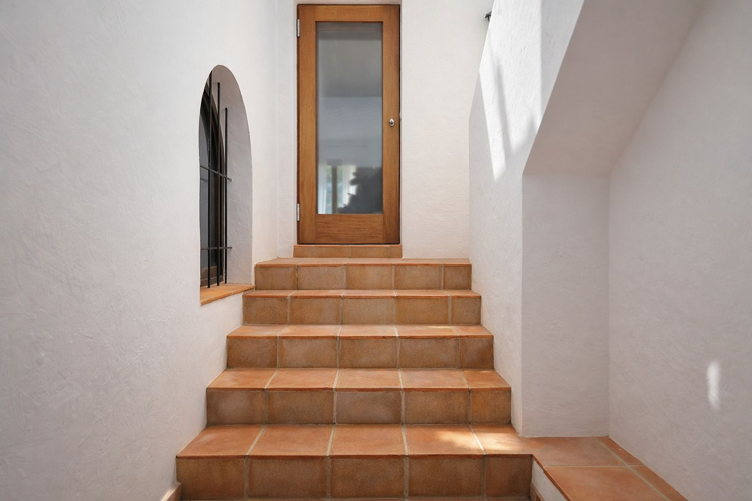 Escalier intérieur aux marches en terre cuite menant à une porte en bois vitrée, agrémenté d'une fenêtre en arche avec ferronnerie et murs blancs.
