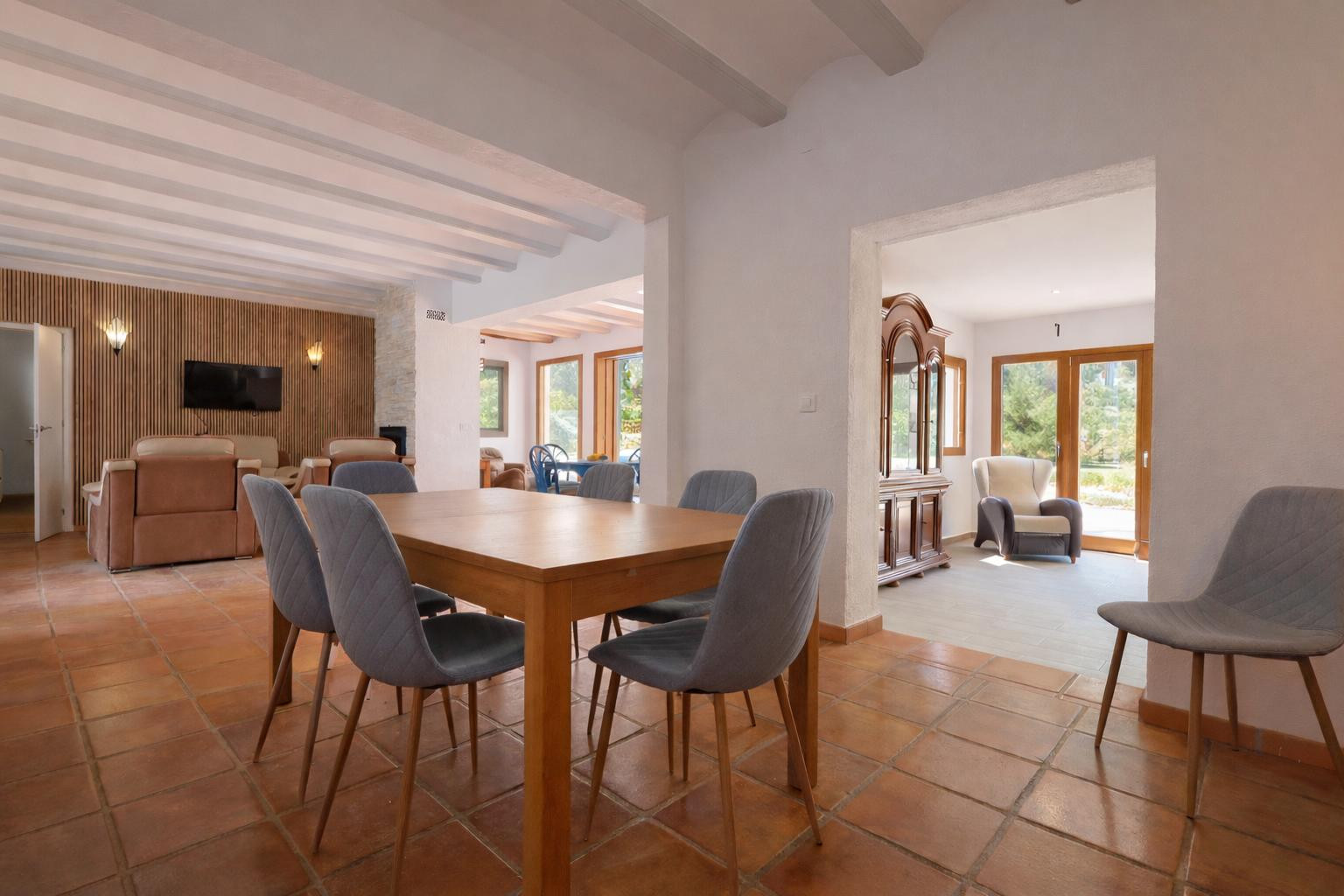 Espace salle à manger avec table en bois, sol en terre cuite et poutres apparentes blanches, ouvert sur le salon et une pièce de vie lumineuse.