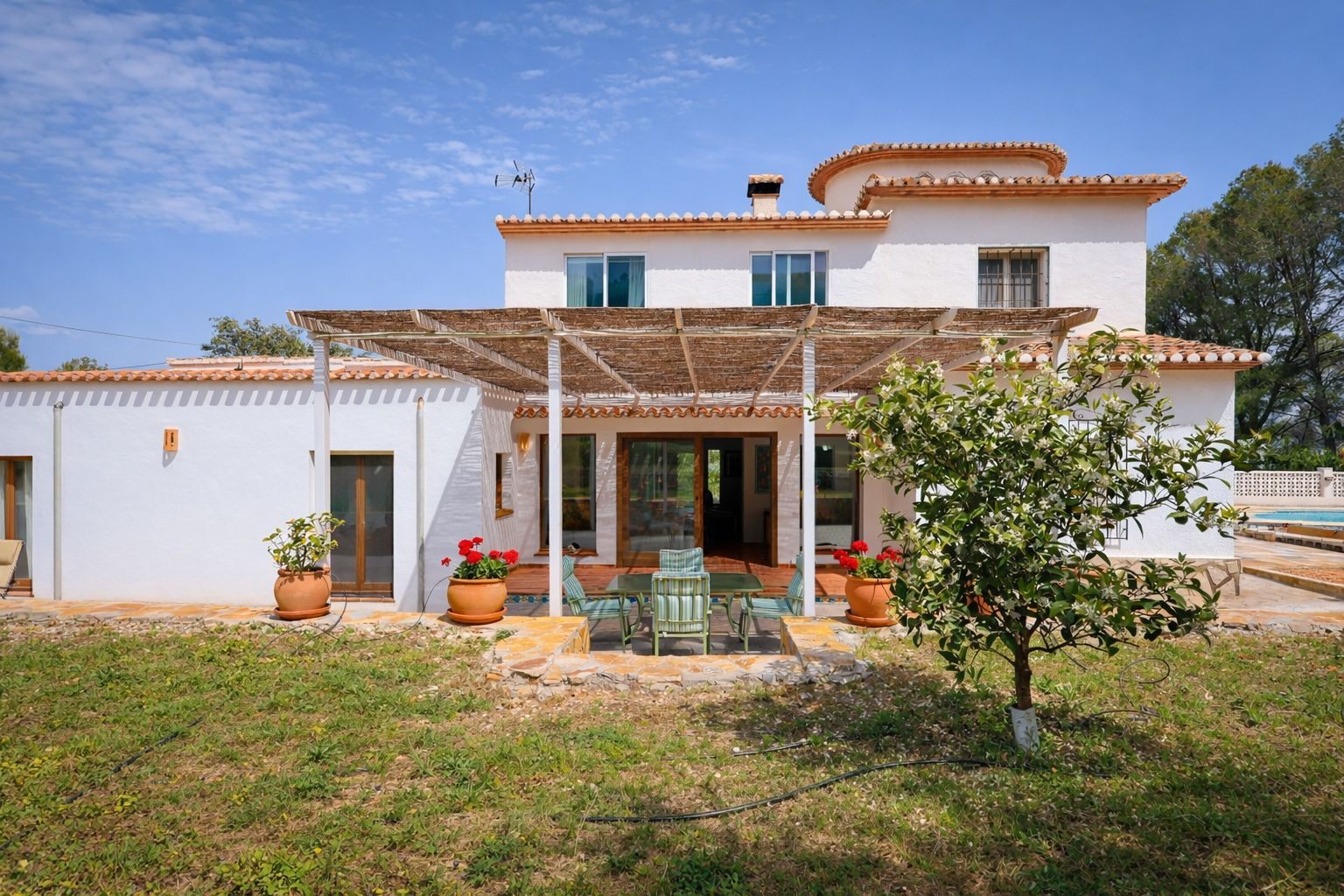Façade arrière de la villa avec pergola en canisse, coin repas extérieur et jardin arboré sous un ciel bleu dégagé.
