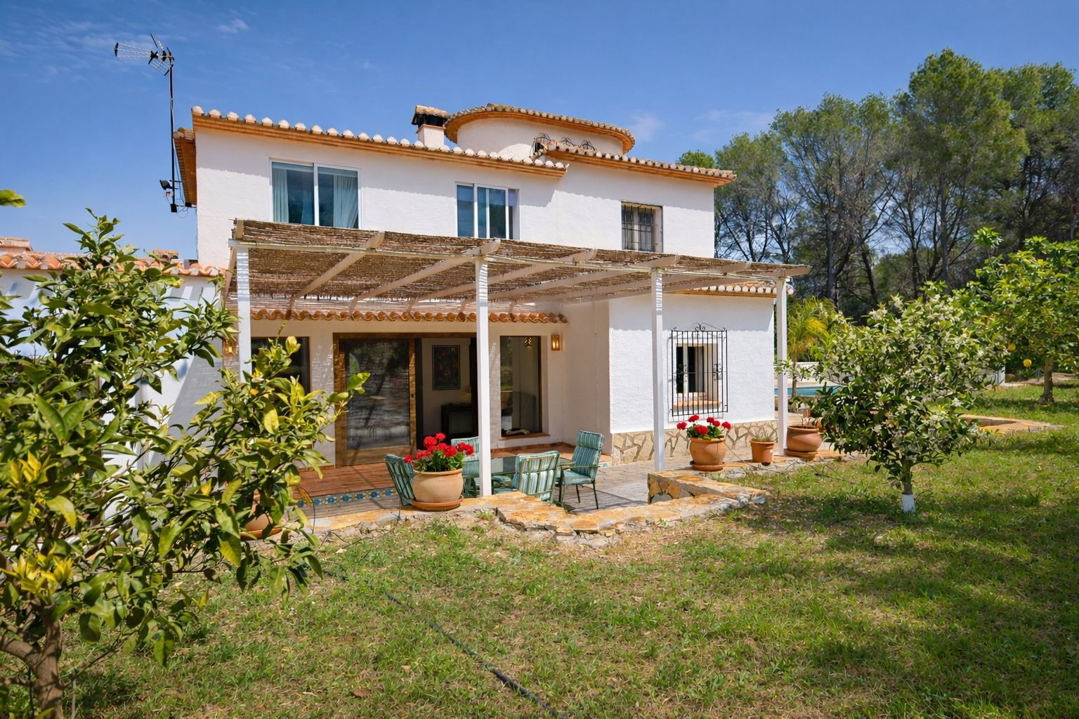 Façade arrière de la villa avec porche en canisse, coin salon extérieur et jardin arboré. L'architecture méditerranéenne présente des détails en pierre et des tuiles romanes.
