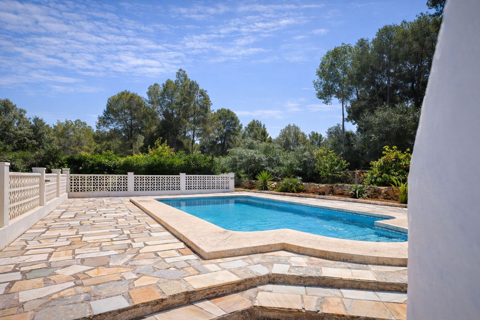 Piscine extérieure avec terrasse en pierre naturelle, clôture décorative blanche et jardin arboré offrant une intimité totale.