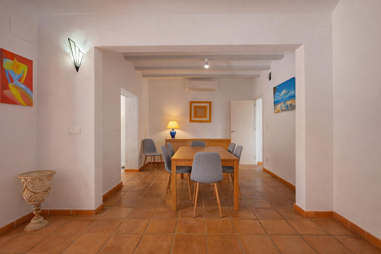 Salle à manger avec table en bois et chaises grises, sol en terre cuite et plafond à poutres apparentes. Équipée d'une unité de climatisation murale.