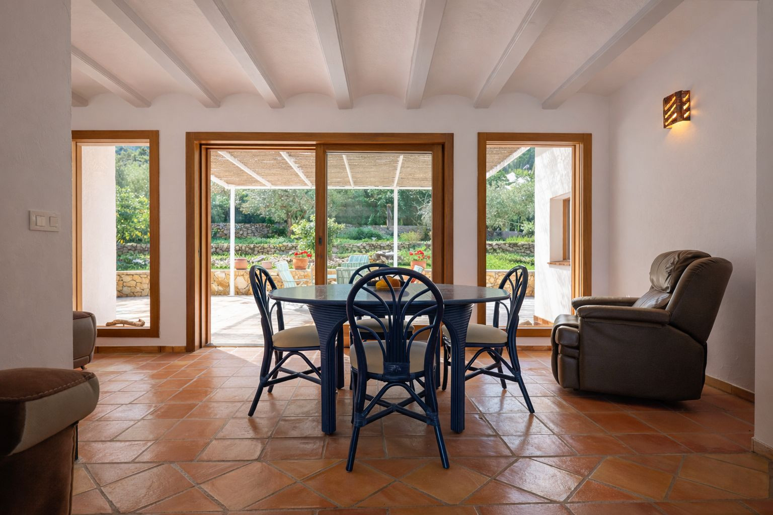 Salle à manger lumineuse avec poutres apparentes blanches, sol en terre cuite et accès direct à la terrasse couverte par de grandes baies vitrées en bois.