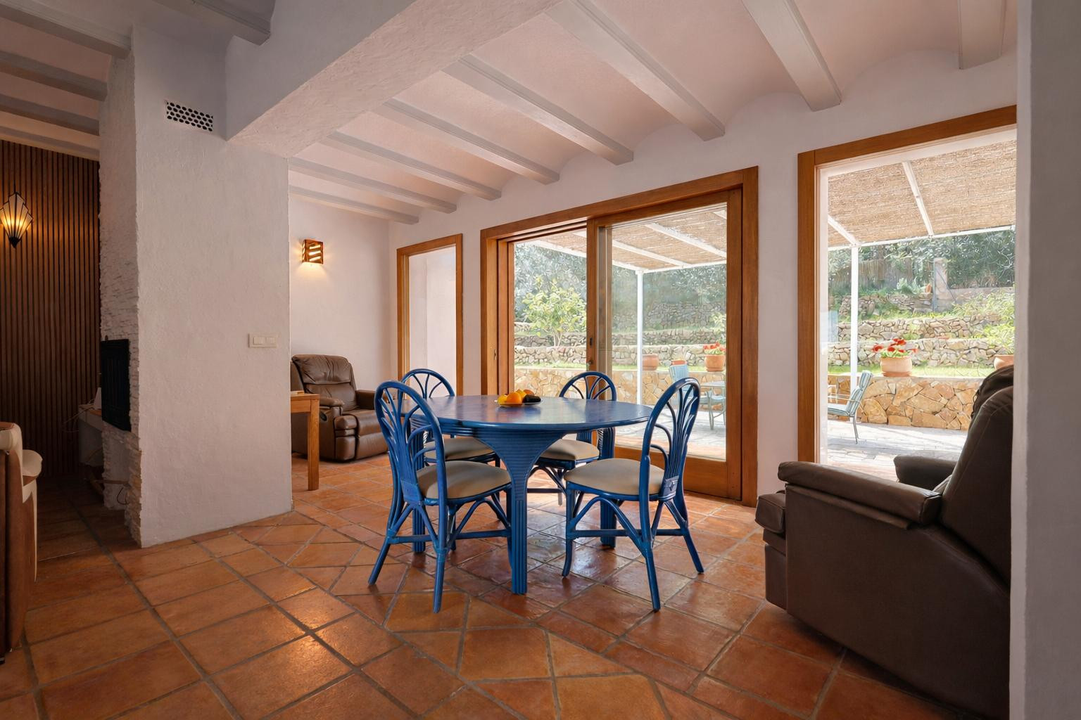 Salle à manger lumineuse avec table bleue, poutres apparentes blanches et sol en terre cuite. De larges baies vitrées en bois s'ouvrent sur le jardin.