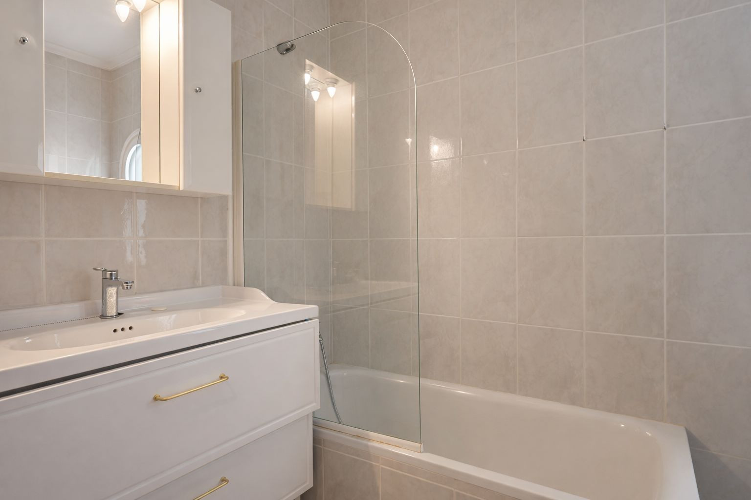 Salle de bain avec baignoire et paroi en verre courbé, meuble vasque blanc aux poignées dorées et armoire de toilette avec éclairage intégré.