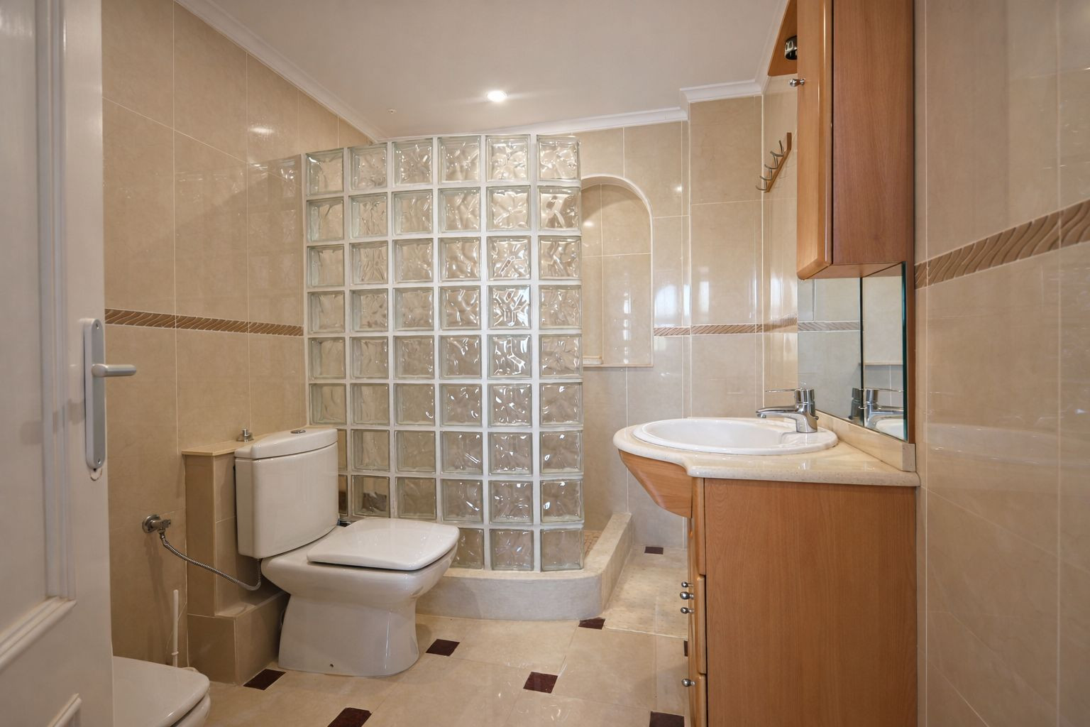 Salle de bain carrelée dans des tons crème, comprenant une douche avec paroi en briques de verre et un meuble vasque en bois avec miroir.
