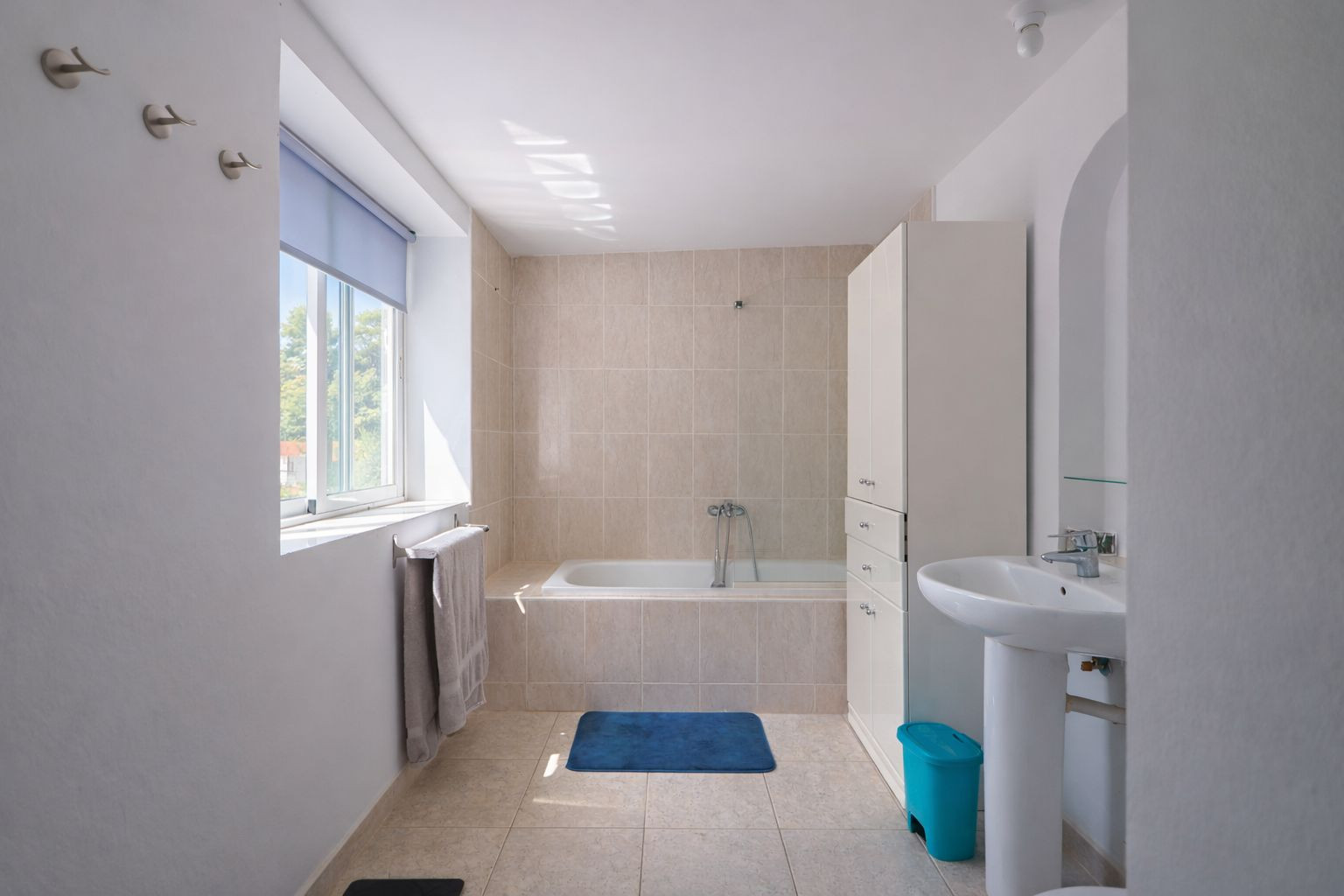 Salle de bain fonctionnelle avec baignoire intégrée, carrelage en tons neutres, lavabo sur colonne et fenêtre lumineuse.