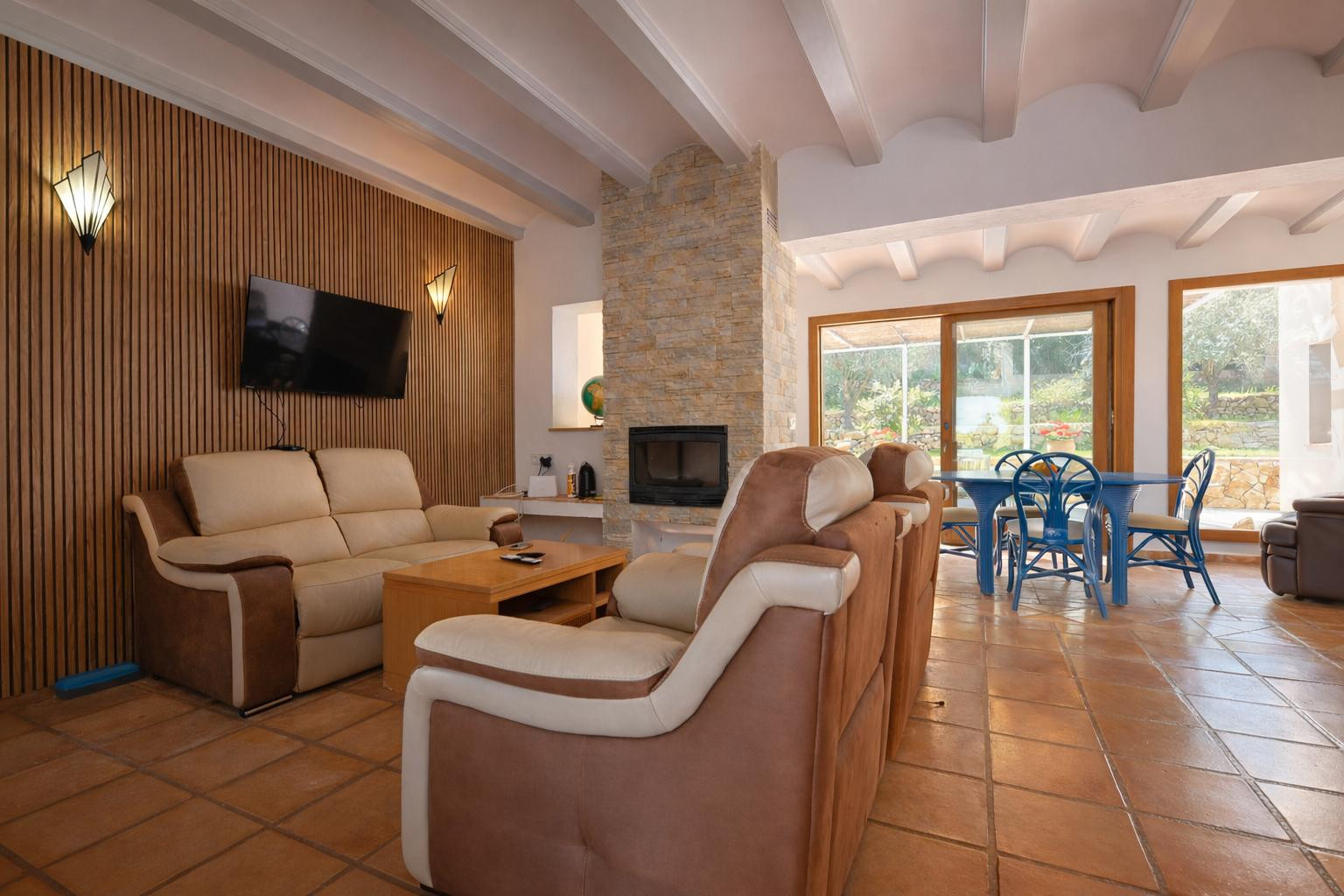Salon-salle à manger avec cheminée en pierre, plafonds à poutres blanches et sol en terre cuite. Comprend un mur en lattes de bois et un accès direct à la terrasse.