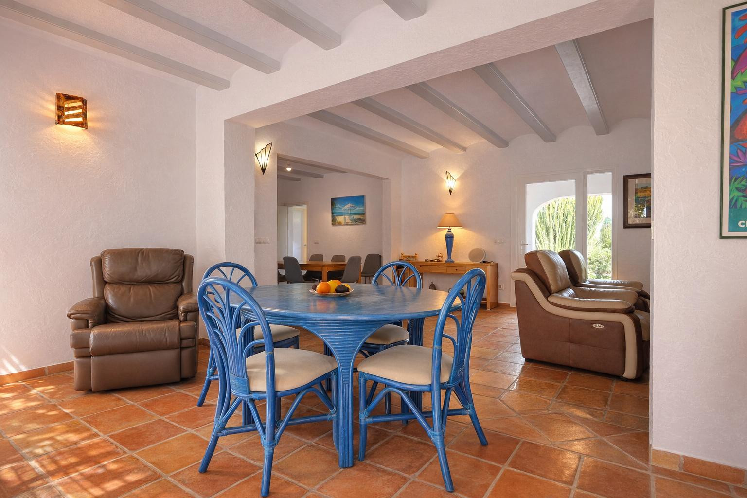 Salon-salle à manger avec sol en terre cuite, poutres apparentes blanches et mobilier en rotin bleu. L'espace ouvert communique avec un second coin salon lumineux.