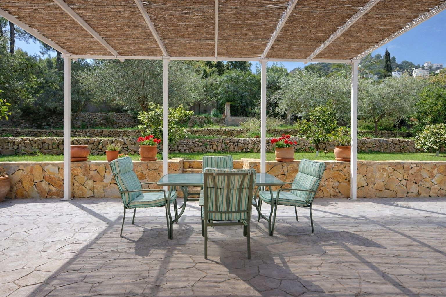 Terrasse extérieure avec pergola en canisse, coin repas et vue sur un jardin en restanques avec murs en pierre naturelle et arbres fruitiers.