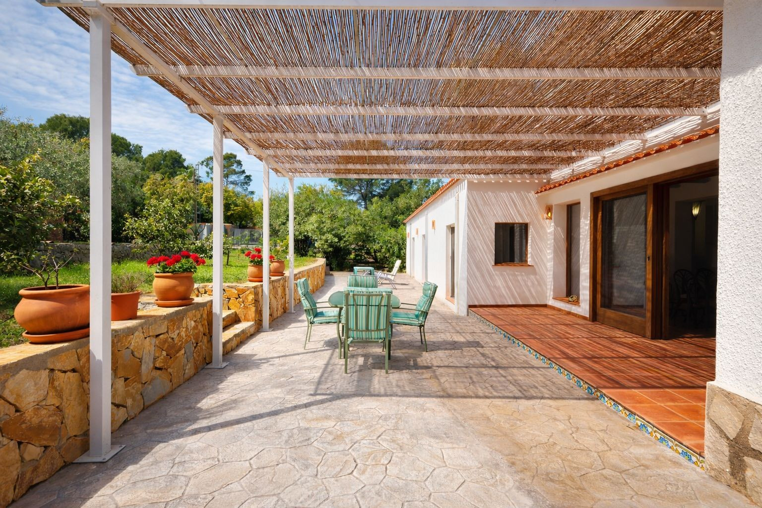 Terrasse extérieure avec pergola en canisse, sol en pierre et mobilier de jardin. Un muret en pierre naturelle avec des pots en terre cuite borde le jardin.