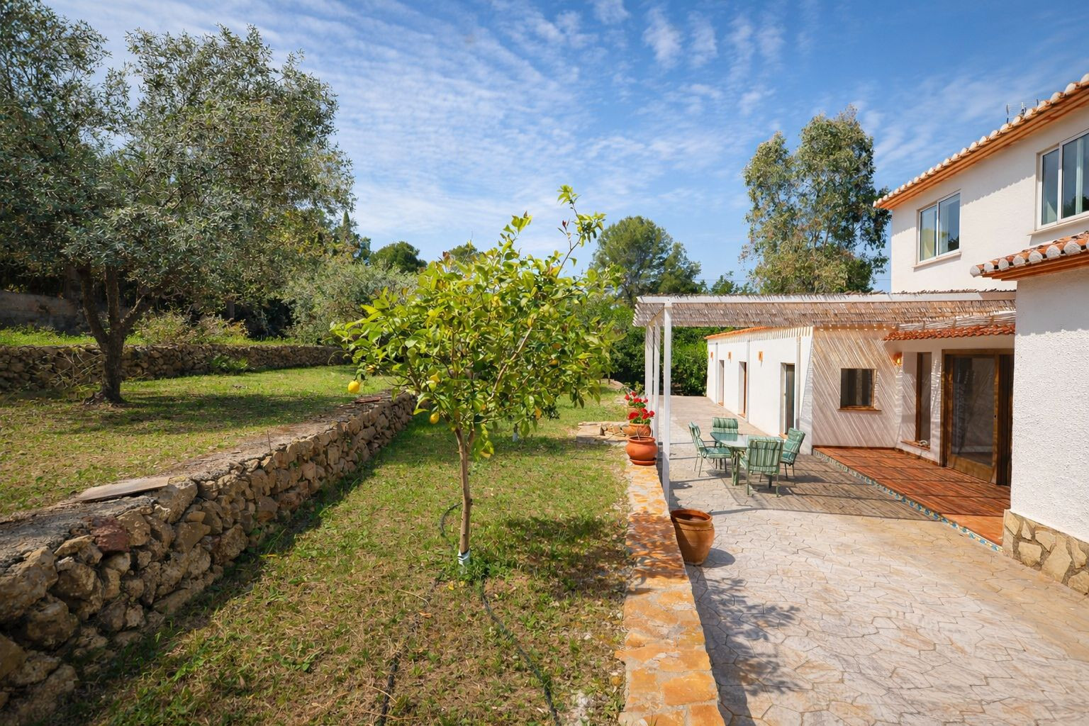 Vue extérieure avec terrasse pavée, pergola ombragée et jardin en restanques avec murs en pierre sèche et arbres fruitiers.