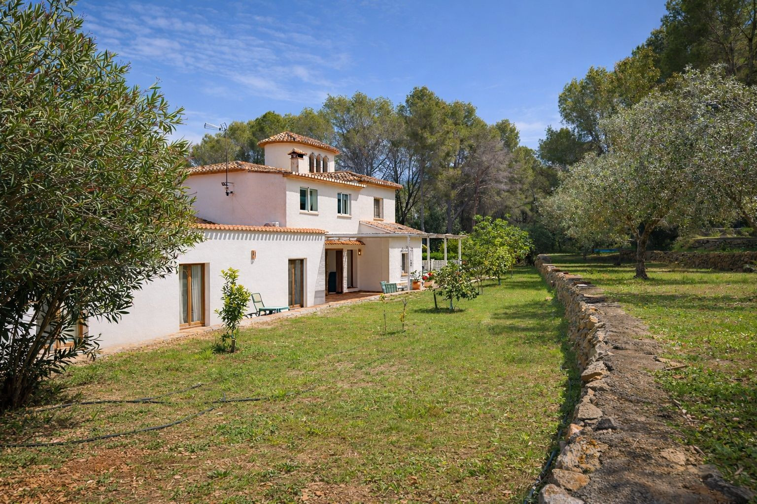 Vue extérieure de la villa à deux étages avec façade blanche, toiture en tuiles, jardin arboré de fruitiers et murets en pierre naturelle.