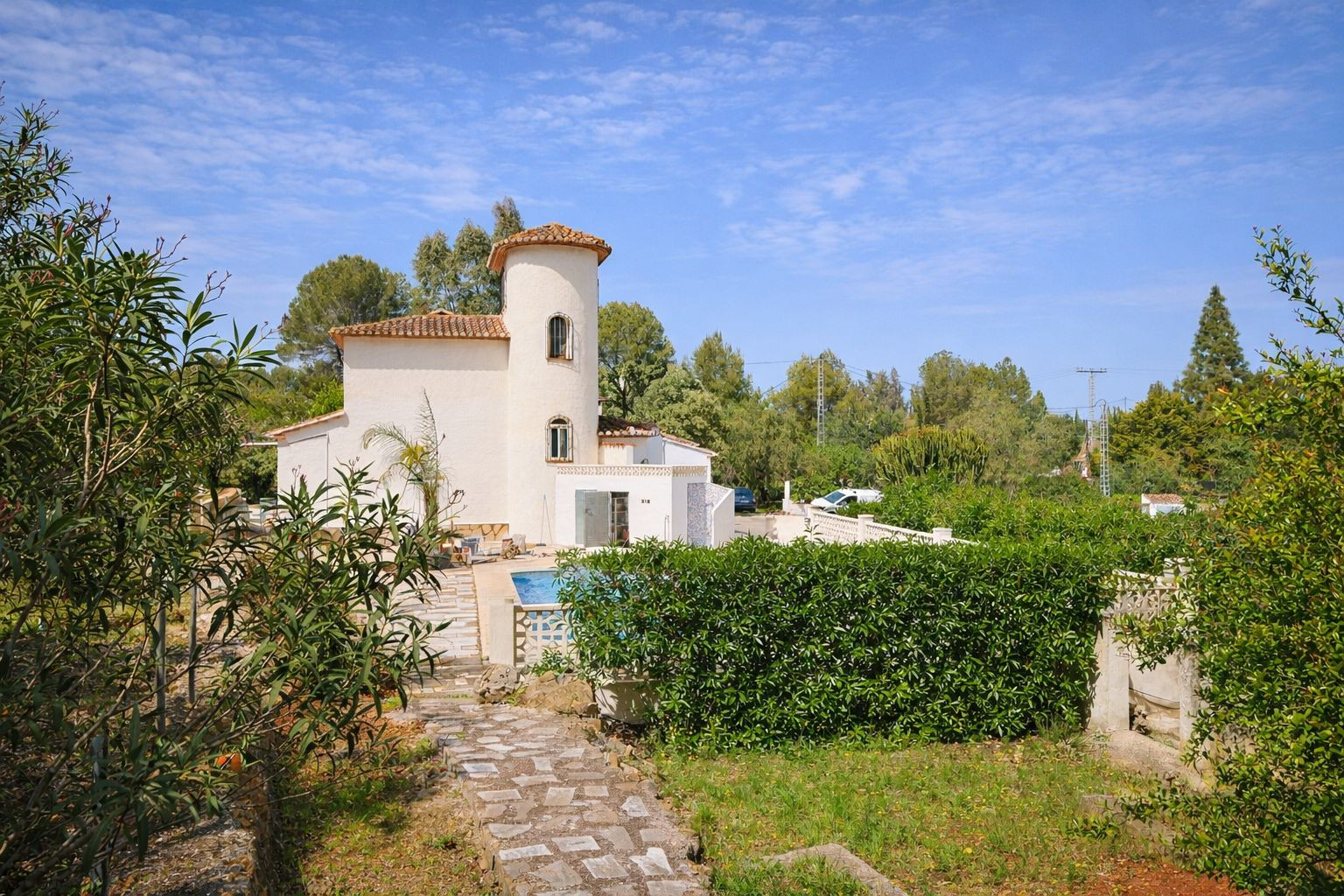Vue extérieure de la villa avec tour circulaire, toit en tuiles romaines et jardin méditerranéen agrémenté de sentiers en pierre et piscine.