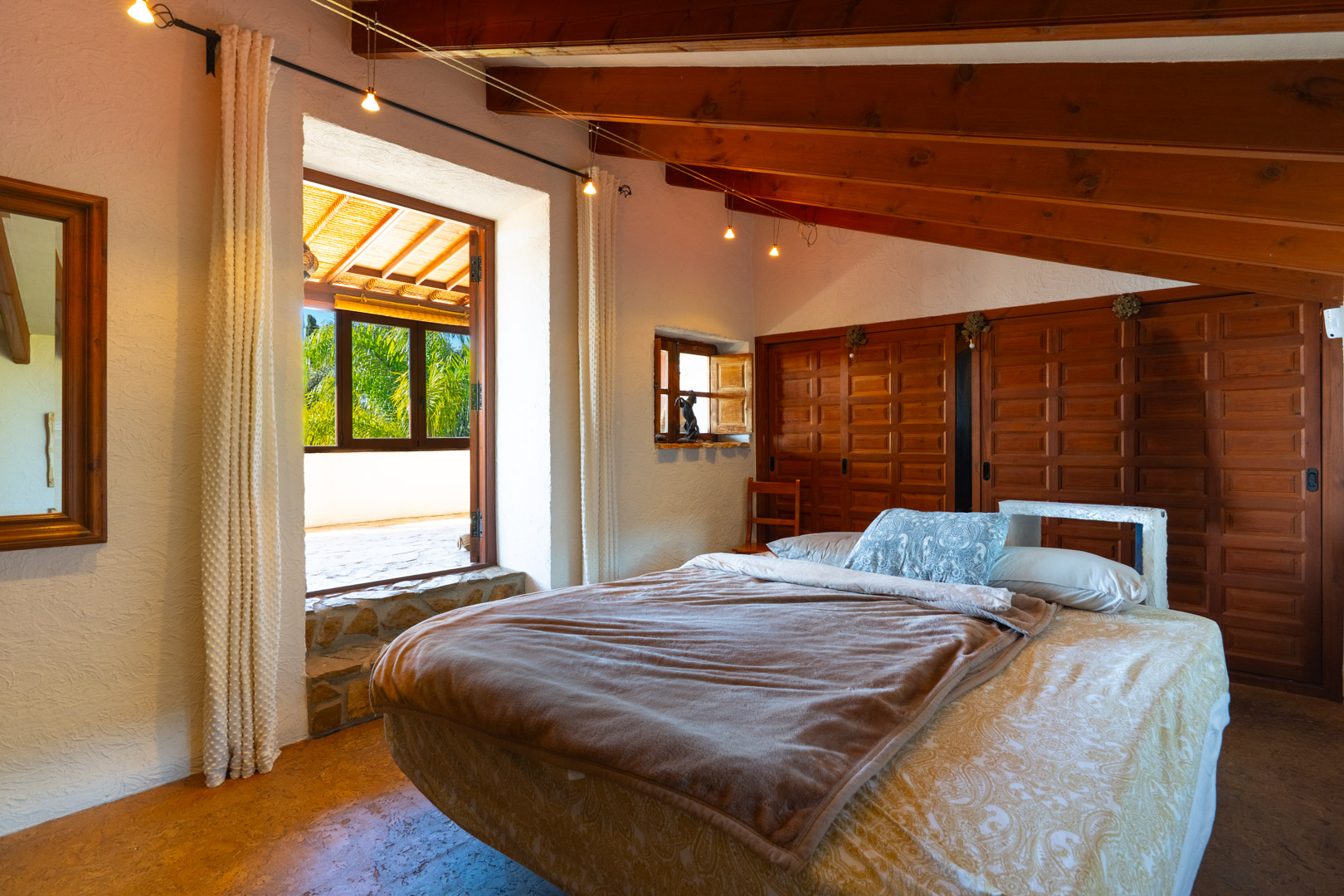 Chambre avec poutres apparentes en bois, placards intégrés et une grande ouverture vers une terrasse privée surplombant le jardin méditerranéen.