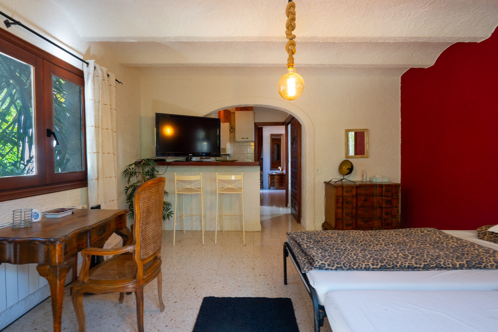 Chambre d'amis avec bureau en bois, mur d'accent rouge et bar avec tabourets ouvrant sur la cuisine par une arche, éclairée par une suspension en corde.