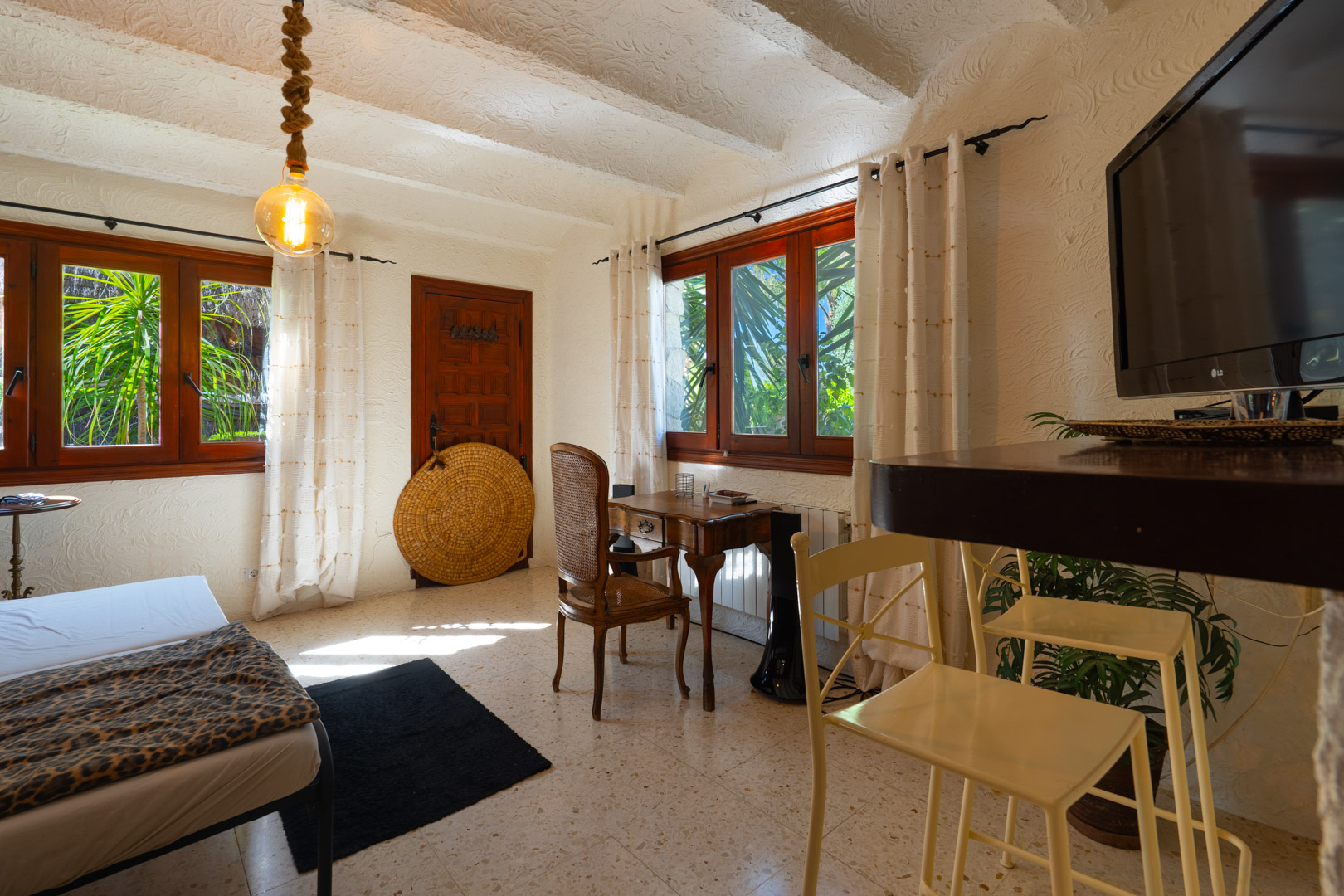 Chambre d'amis rustique avec plafonds à poutres blanches, bureau en bois classique et fenêtres à cadre sombre offrant une vue sur la végétation.