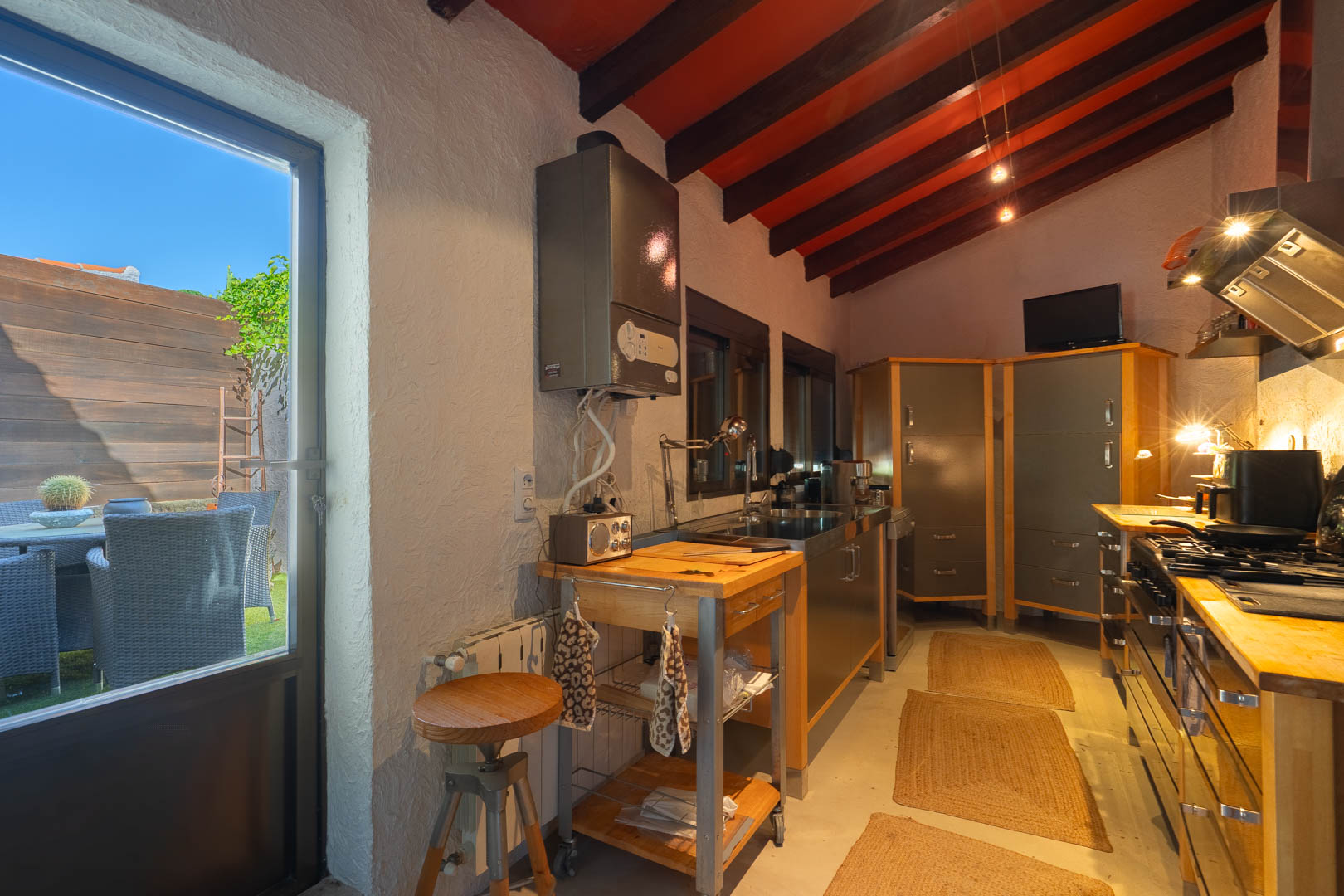 Cuisine rustique avec poutres en bois sombres, îlot de travail mobile et accès direct à un patio extérieur privé par une porte vitrée.