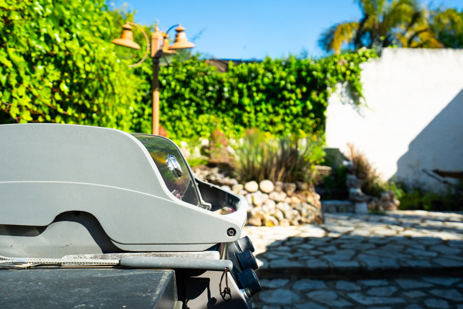 Espace barbecue extérieur situé sur un patio en pierre naturelle, entouré d'une végétation méditerranéenne luxuriante et d'un lampadaire décoratif.
