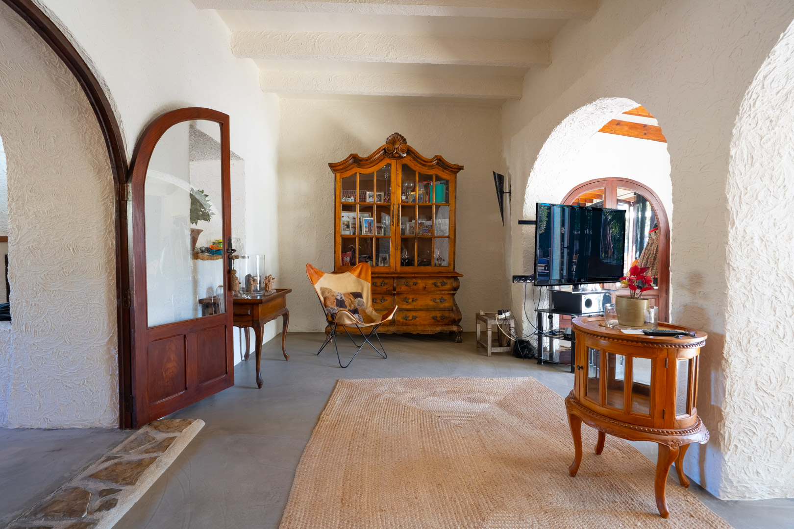Espace de vie avec plafonds à poutres blanches, arches traditionnelles et sol en microciment. Comprend une vitrine ancienne en bois et du mobilier rustique.