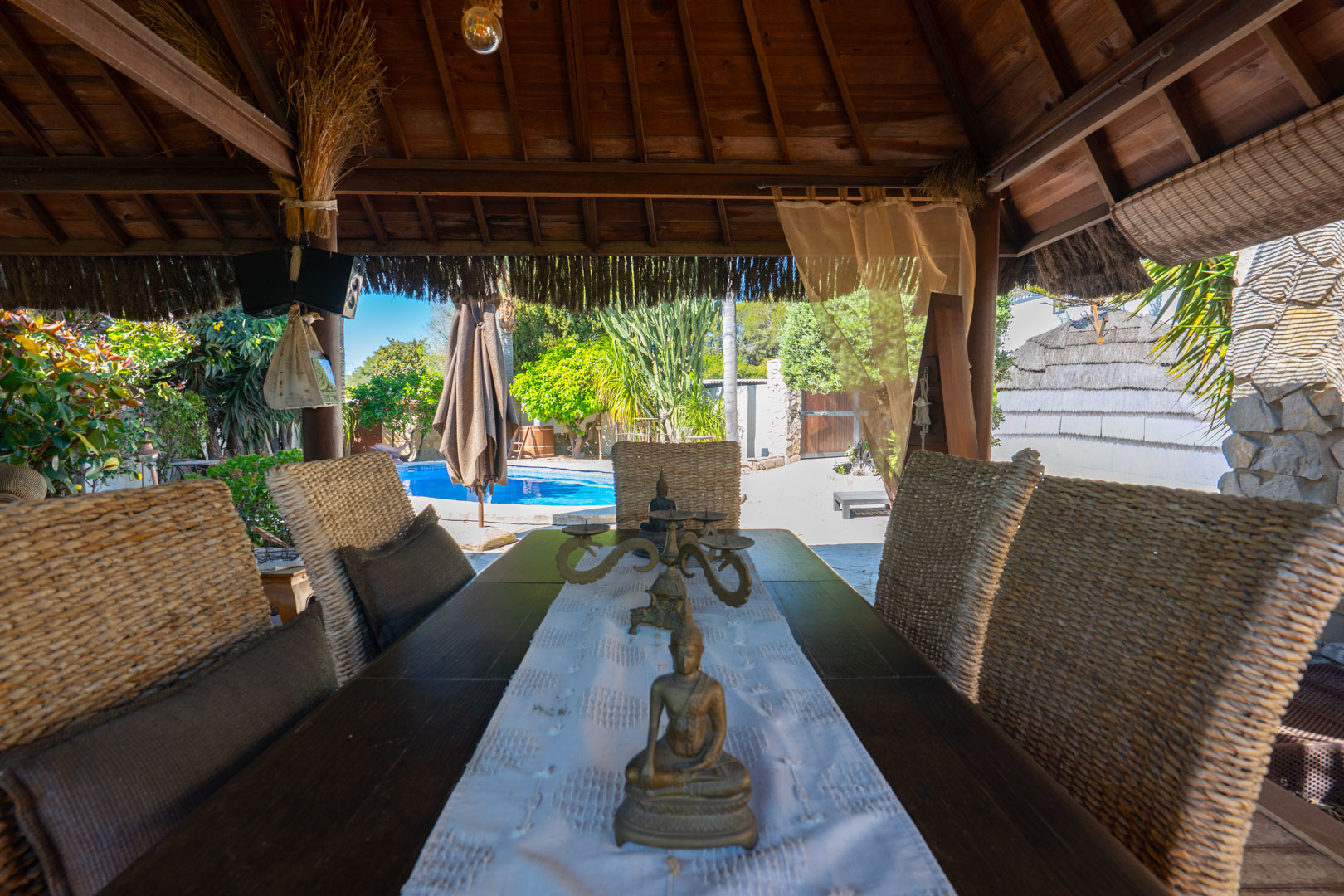 Espace dînatoire extérieur sous une pergola balinaise en bois, avec chaises en osier et vue sur la piscine et le jardin méditerranéen.