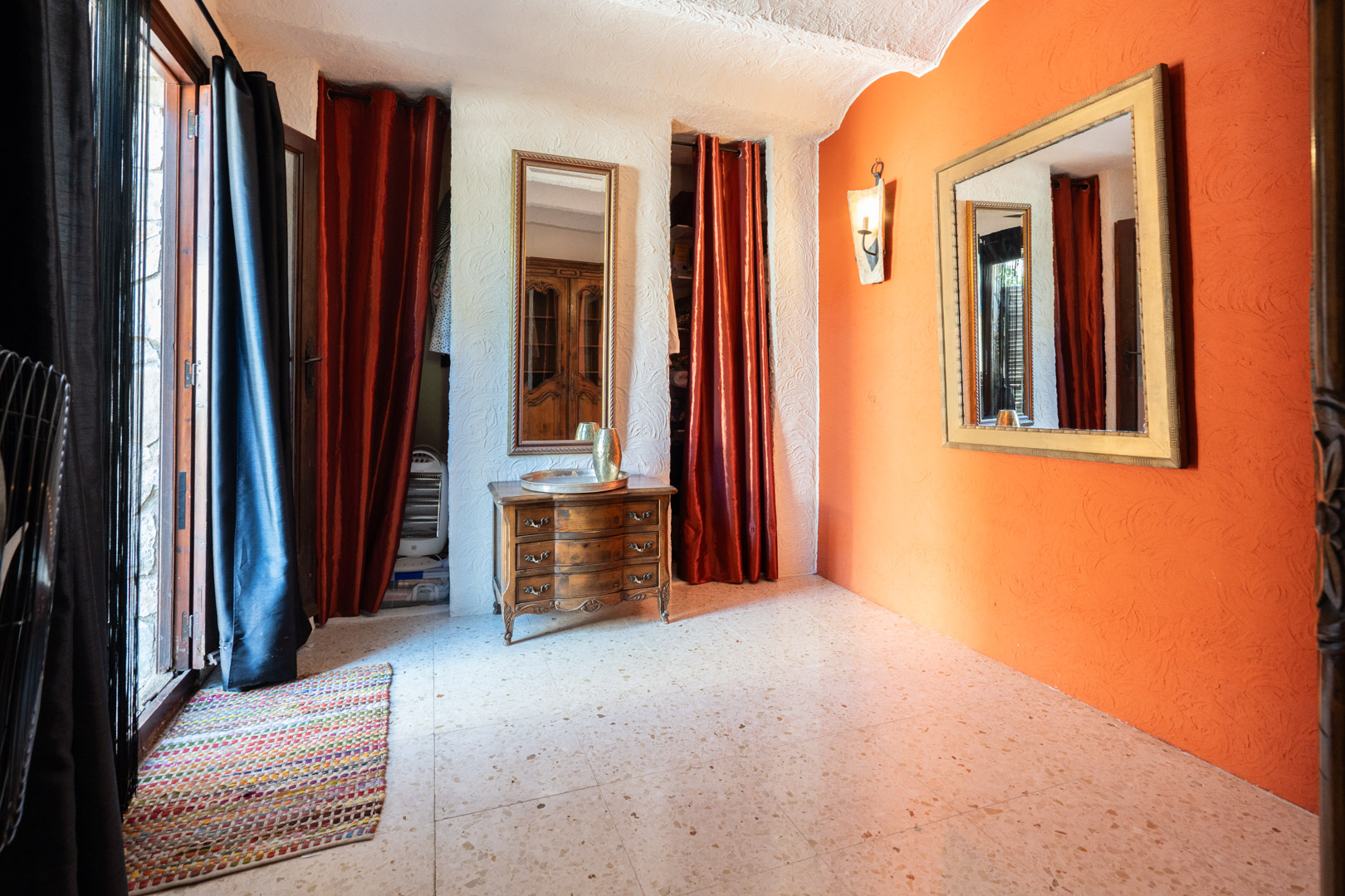 Espace dressing avec mur d'accent orange, miroirs ornés, commode en bois classique et sol en terrazzo. Des rideaux rouges dissimulent les espaces de rangement intégrés.