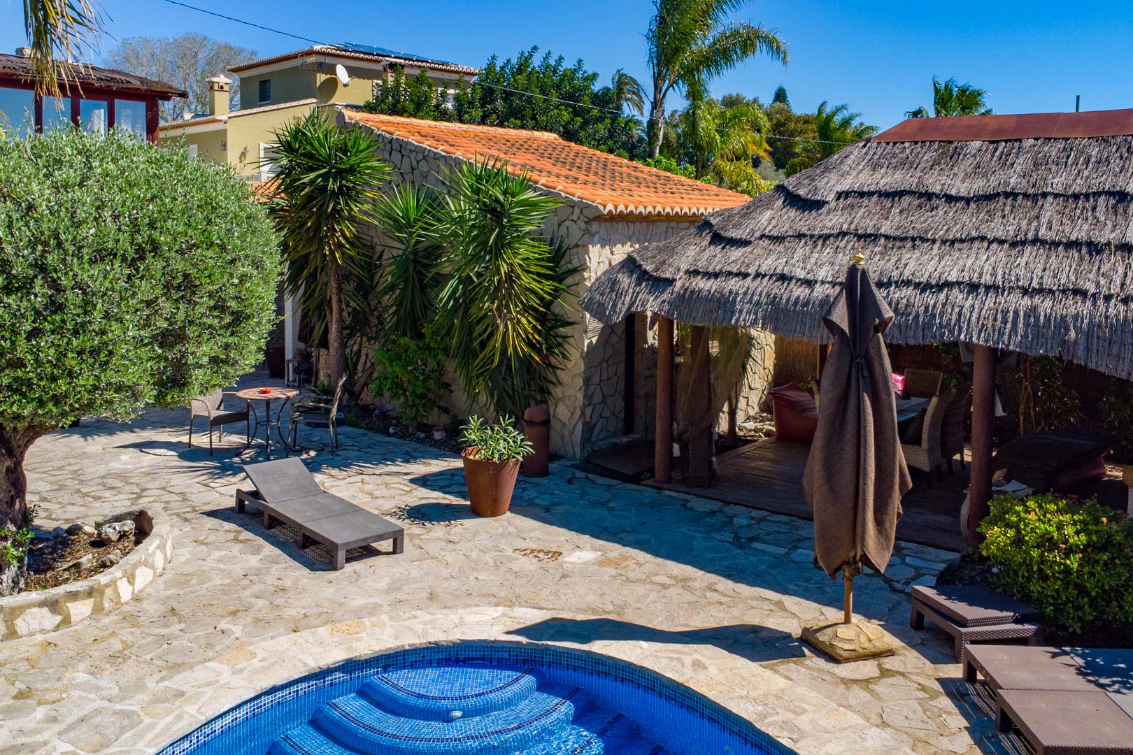 Espace extérieur avec terrasse en pierre naturelle, piscine et pergola balinaise en chaume, entouré d'une bâtisse en pierre et d'une végétation méditerranéenne.