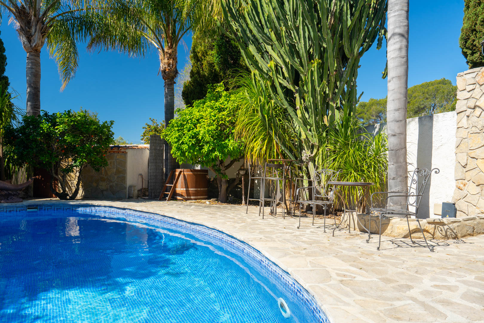 Espace piscine avec terrasse en pierre naturelle, mobilier en fer forgé et jardin méditerranéen composé de grands cactus et de palmiers.