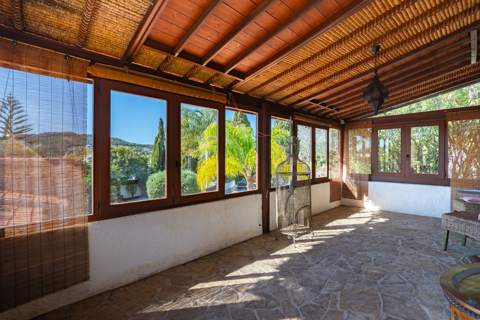 Grande naya vitrée avec sol en pierre naturelle, plafond en poutres de bois et canisse, offrant une vue dégagée sur le jardin méditerranéen et les collines.