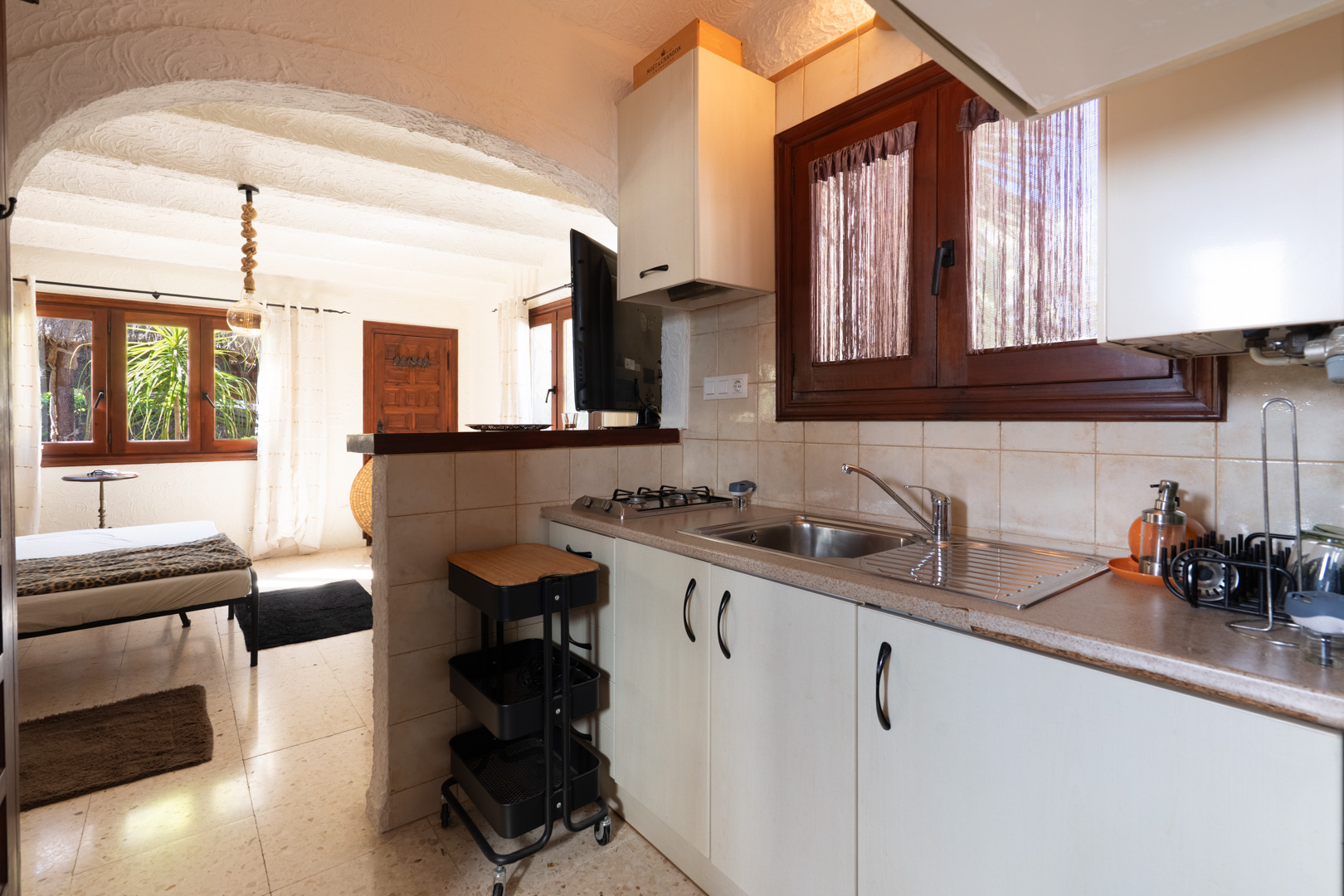 Kitchenette rustique avec meubles blancs et menuiseries en bois, s'ouvrant par une arche sur une chambre lumineuse au sol carrelé.