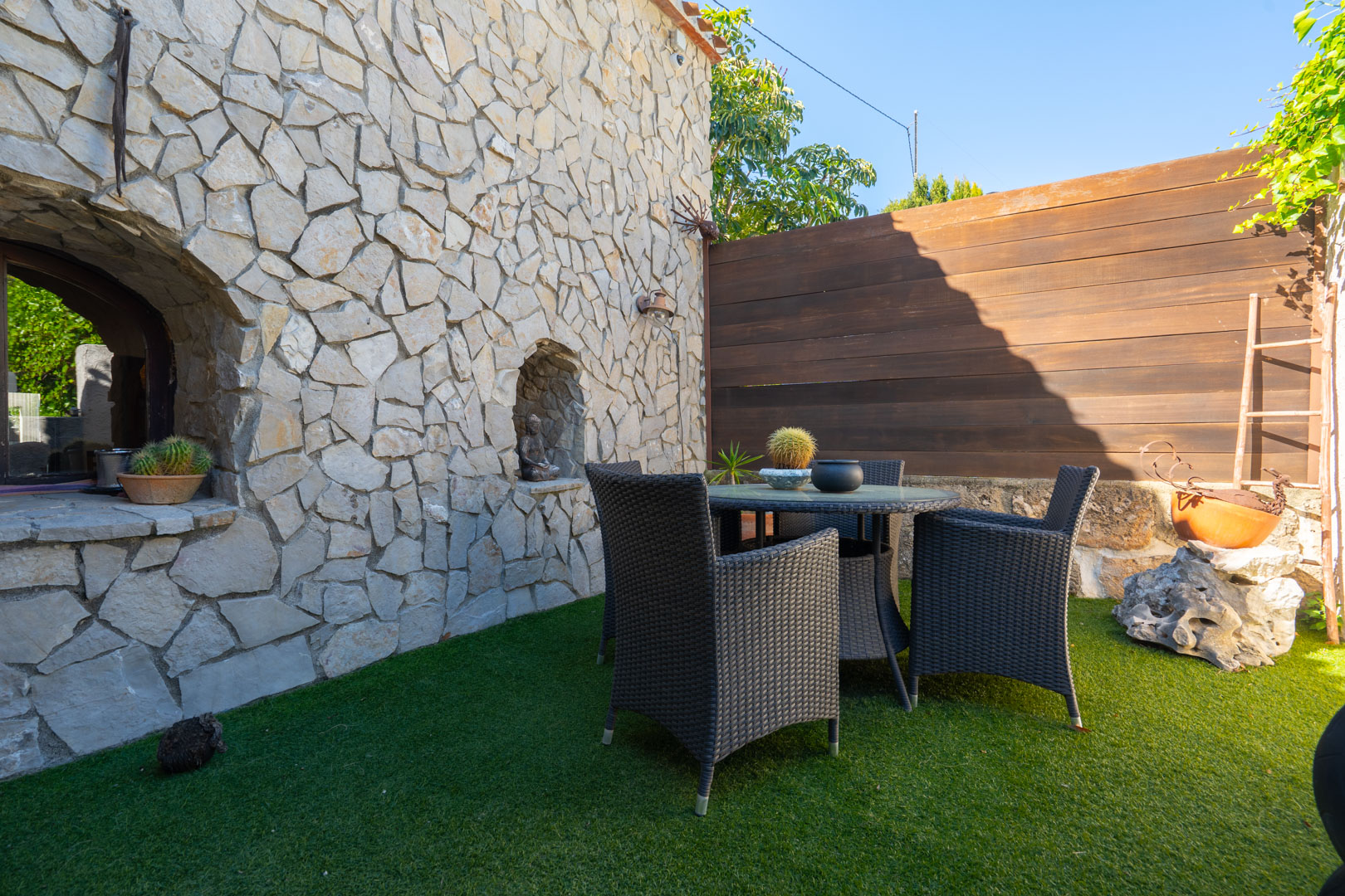 Patio extérieur doté d'un mur en pierre naturelle avec niches décoratives, un ensemble de repas en rotin sombre et un revêtement en gazon synthétique.