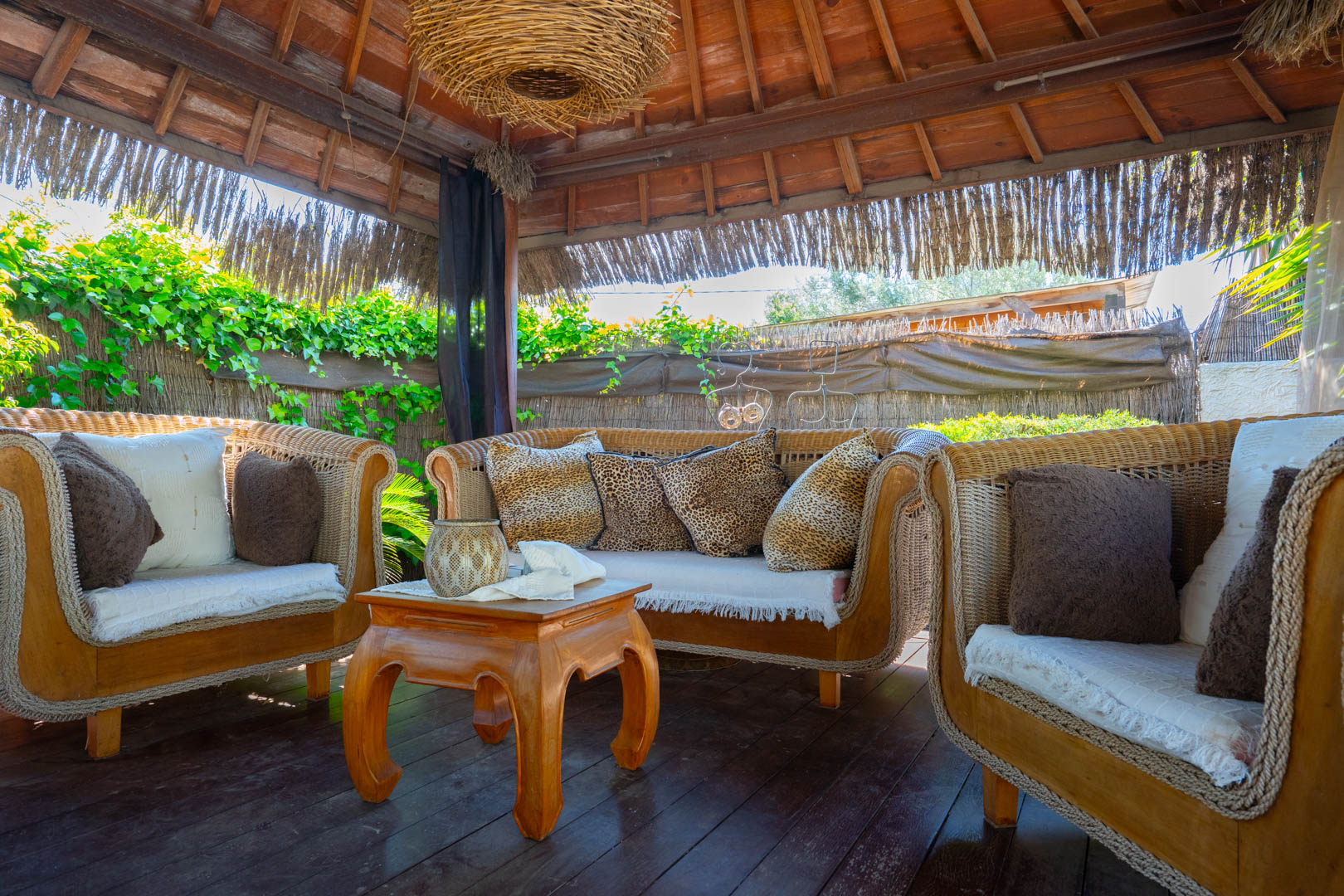 Pergola balinaise avec plafond en bois apparent, mobilier de salon en osier et table basse artisanale sur un sol en terrasse boisée.