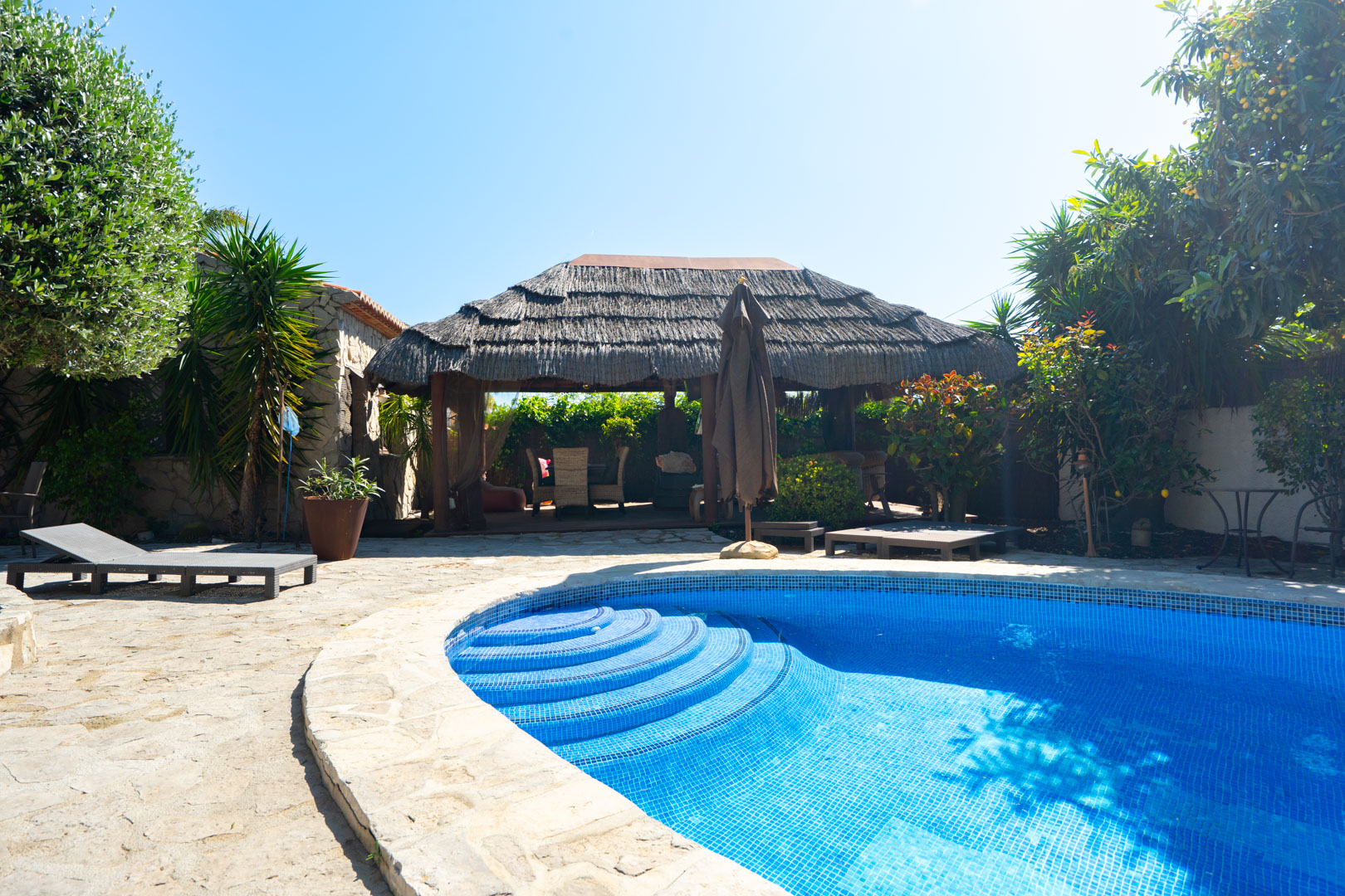 Piscine extérieure avec mosaïque bleue et escalier roman, entourée d'une terrasse en pierre et d'une pergola balinaise au toit de chaume.