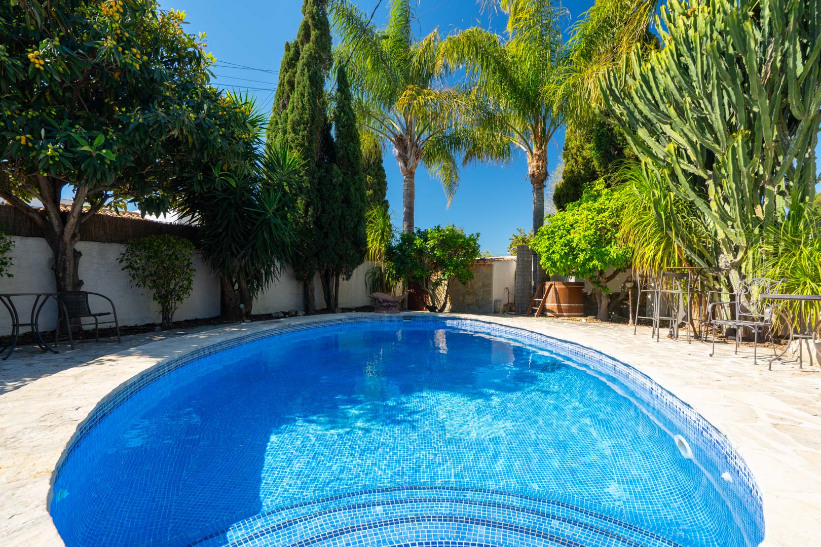 Piscine ovale aux mosaïques bleues, entourée d'une terrasse en pierre naturelle et d'un jardin méditerranéen luxuriant avec palmiers et cyprès.