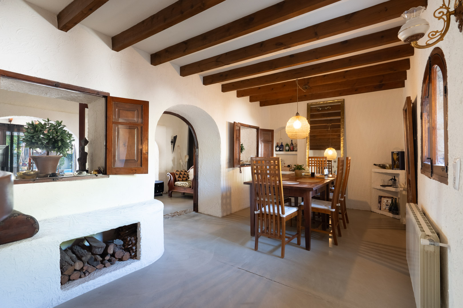 Salle à manger rustique avec poutres apparentes, table en bois massif et arche traditionnelle. Comprend un rangement pour bois intégré et des volets intérieurs en bois.