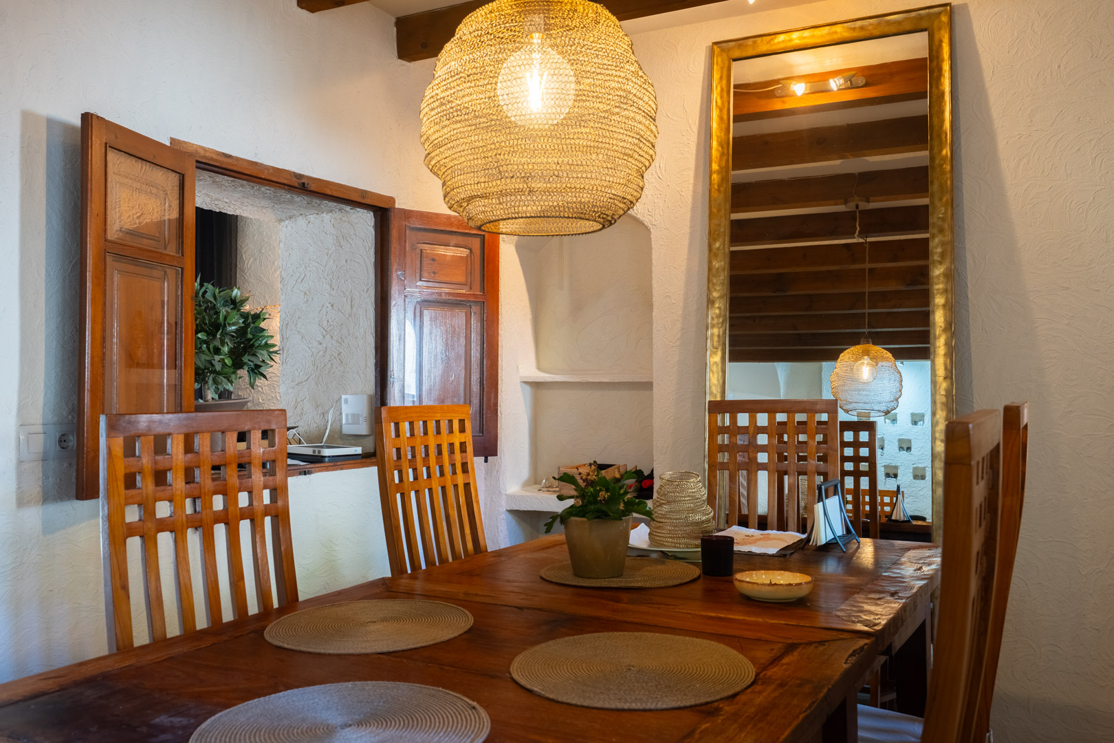 Salle à manger rustique avec table en bois massif, chaises à haut dossier et suspension en fibres naturelles. Des poutres apparentes et un grand miroir doré complètent l'espace.