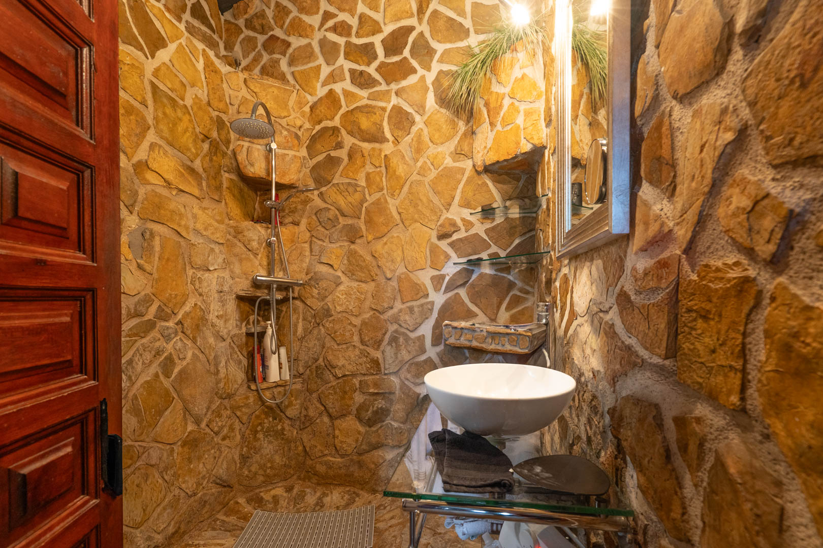 Salle de bain rustique avec murs en pierre naturelle, douche à l'italienne avec étagères intégrées et vasque à poser sur support en verre.