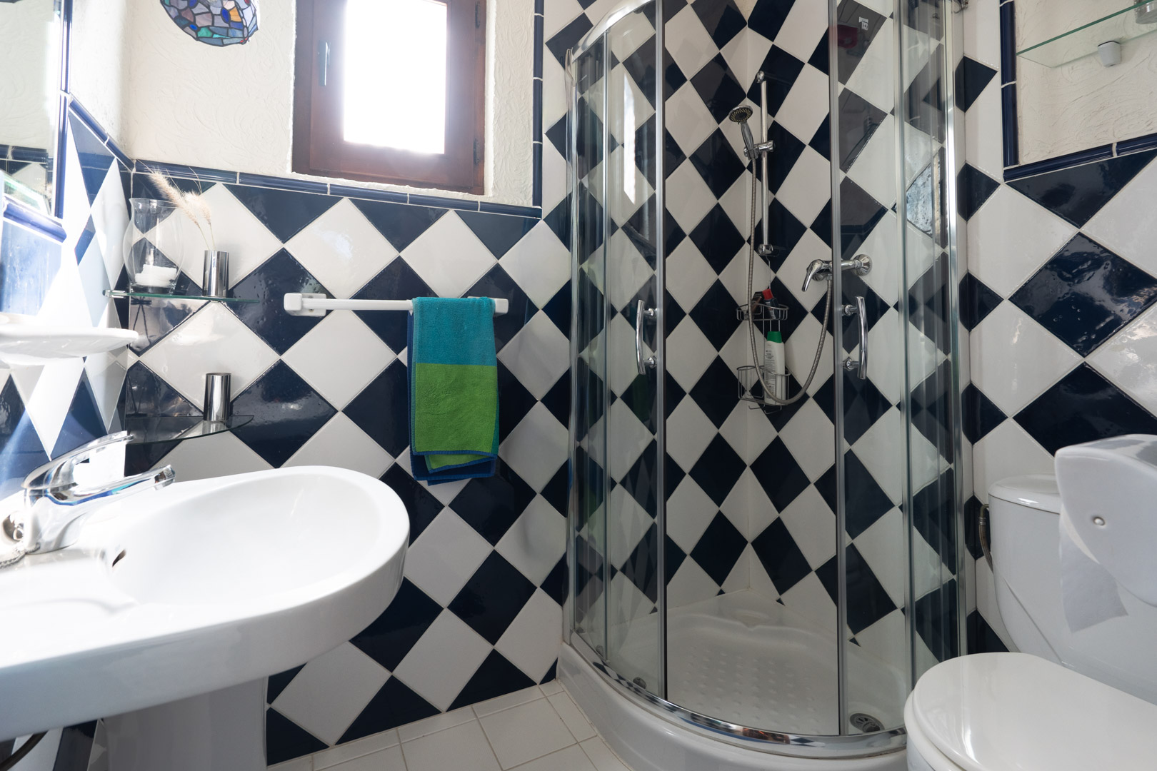 Salle de bains avec carrelage en damier bleu et blanc, comprenant une cabine de douche d'angle vitrée, un lavabo sur colonne et une petite fenêtre en bois.