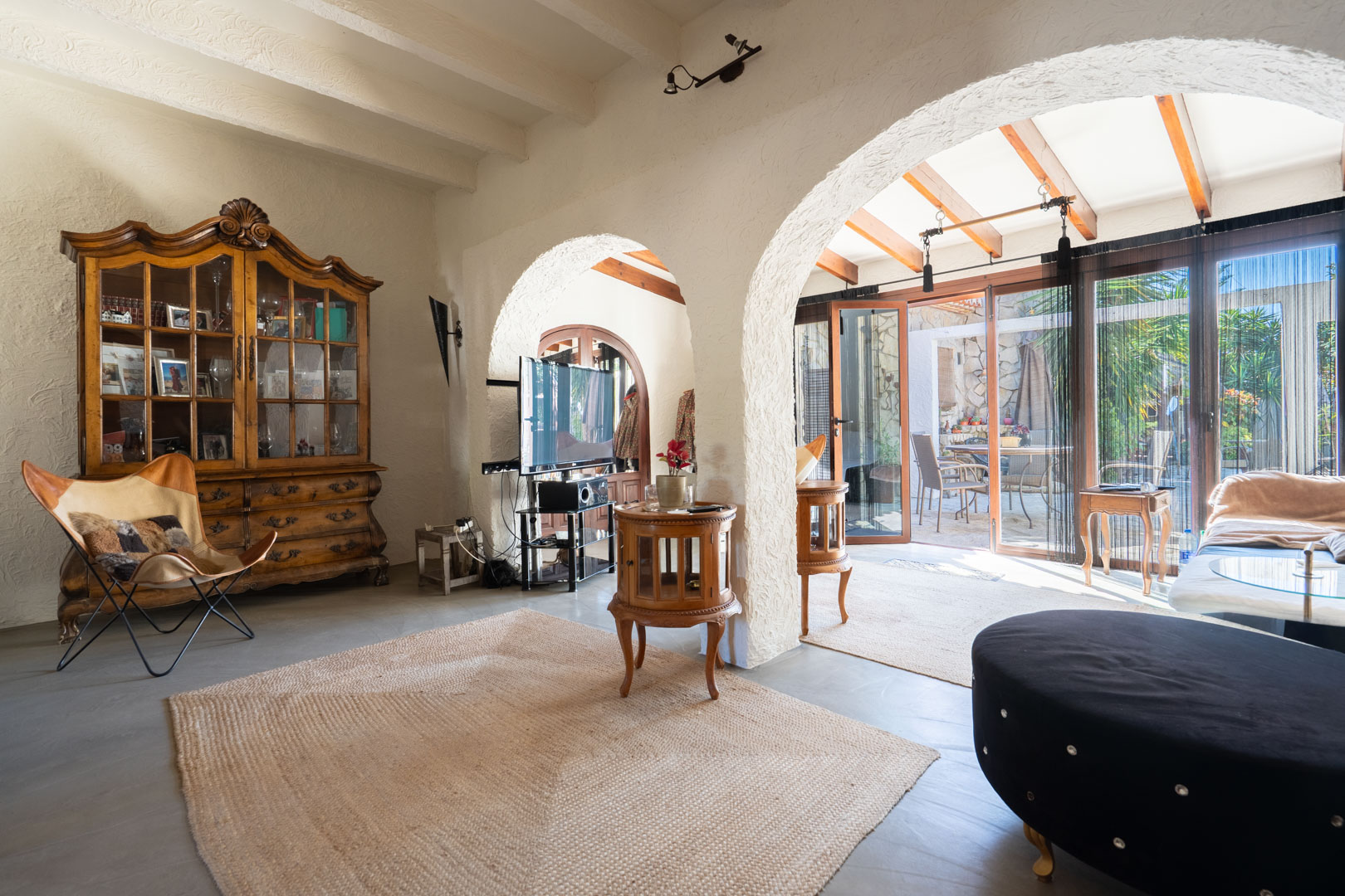 Salon rustique avec plafonds à poutres blanches et arches ouvrant sur une naya. Comprend une vitrine ancienne en bois, un tapis en jute et une lumière naturelle traversante.