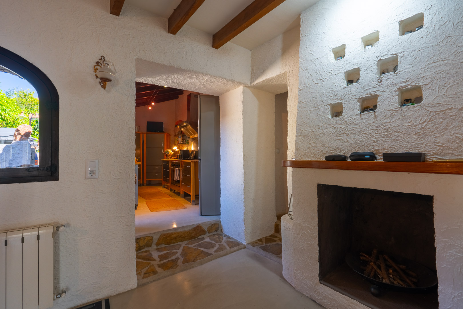 Salon rustique avec poutres en bois, cheminée maçonnée avec niches décoratives et passage vers la cuisine souligné par un sol en pierre naturelle.