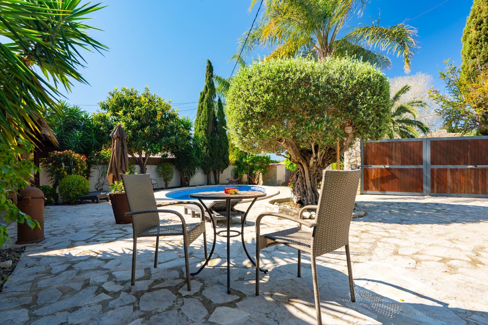 Terrasse extérieure avec pavage en pierre naturelle, mobilier en rotin et vue sur la piscine privée entourée d'oliviers et d'une végétation méditerranéenne luxuriante.