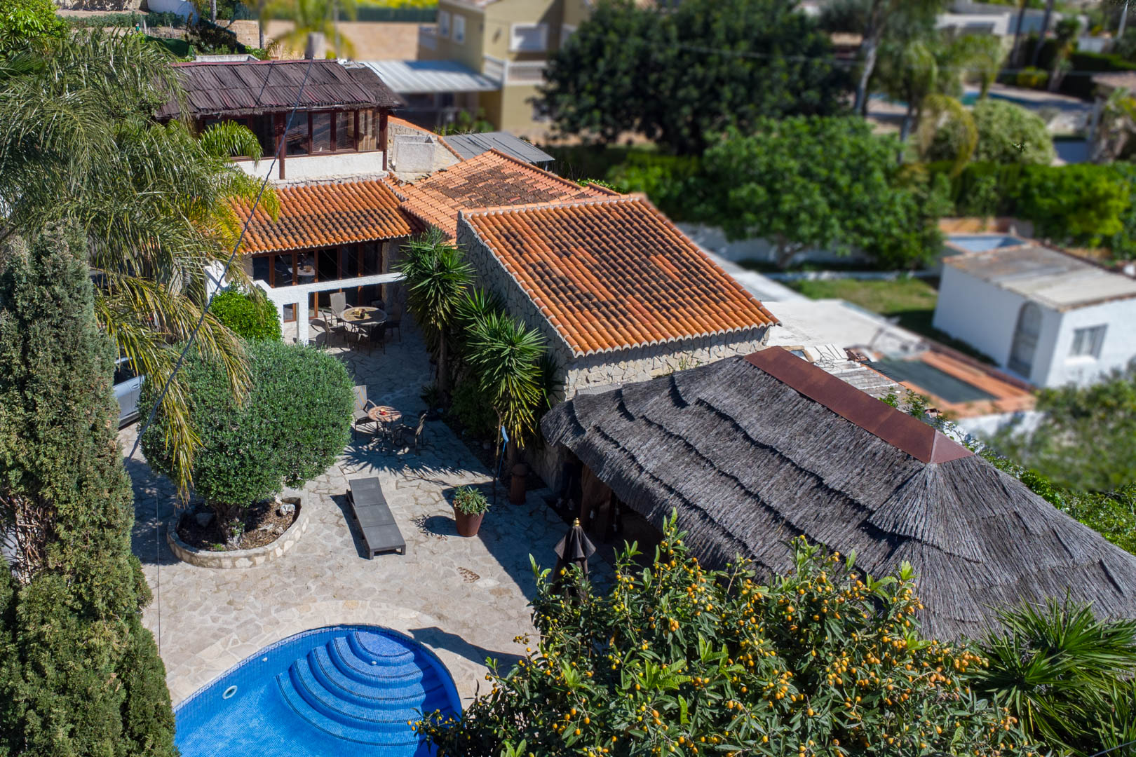 Vue aérienne de la villa avec toits en terre cuite, piscine circulaire en mosaïque bleue, vastes terrasses en pierre et une grande pergola balinaise en chaume.