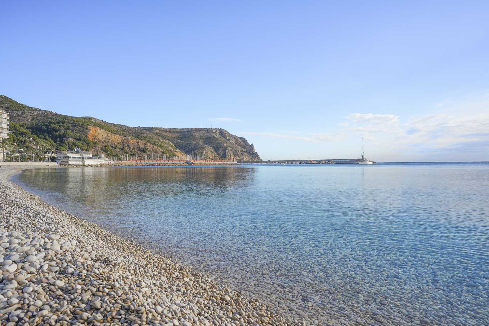 Vue de la plage de galets aux eaux cristallines avec le port en arrière-plan, dominée par les falaises du Cabo de San Antonio à Jávea.
